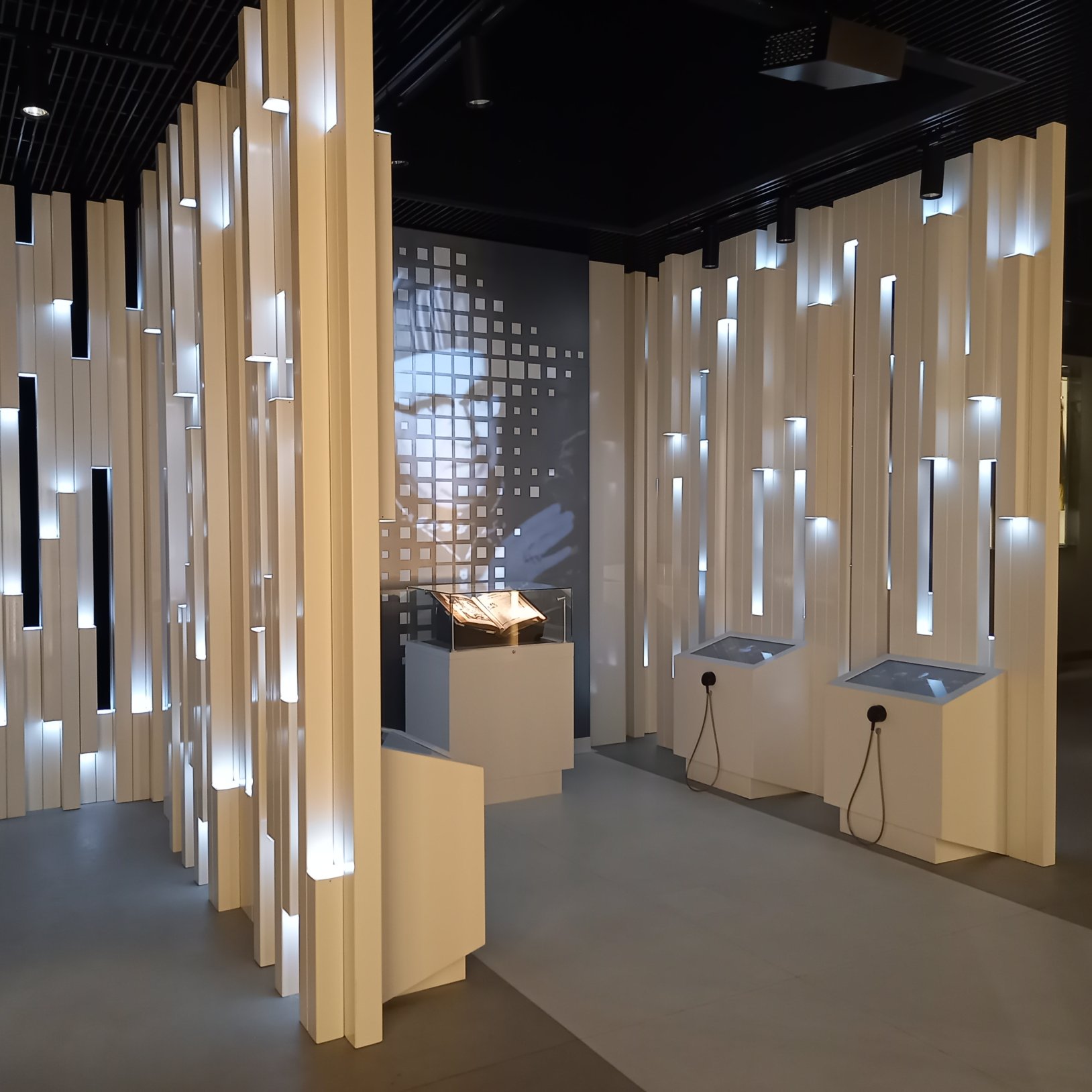 "Nowoczesna sala wystawowa z podświetlanymi białymi panelami ściennymi i ekranami dotykowymi. W centralnym punkcie ekspozycji znajduje się zabytkowa księga umieszczona w szklanej gablocie.