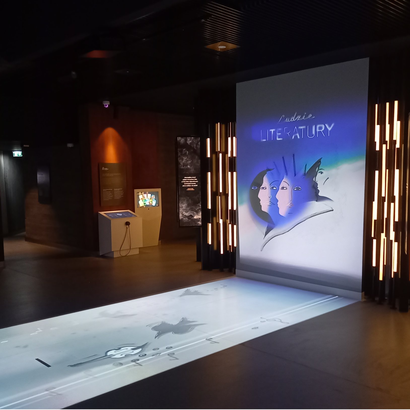 "Interaktywna część wystawy muzealnej z podłogową projekcją i ekranem przedstawiającym grafikę z napisem 'Ludzie Literatury'. Na ilustracji widoczne są stylizowane twarze kobiet. Otoczenie jest ciemne, z podświetlonymi elementami wystroju po bokach."