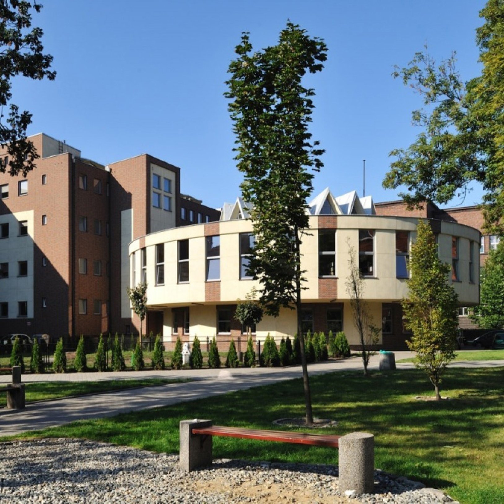 "Nowoczesny budynek o okrągłej bryle, otoczony zielenią. Elewacja łączy jasny tynk i cegłę. Na pierwszym planie znajdują się drzewa, alejka i ławka w parku."