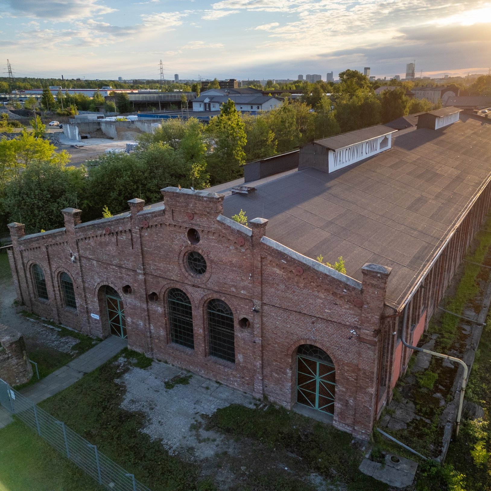 "Widok z góry na ceglaną halę przemysłową o długim dachu; na szczycie budynku widoczny napis „WALCOWNIA CYNKU”, wokół rosną drzewa, a w tle widać zabudowania i panoramę miasta w ciepłym świetle zachodzącego słońca."
