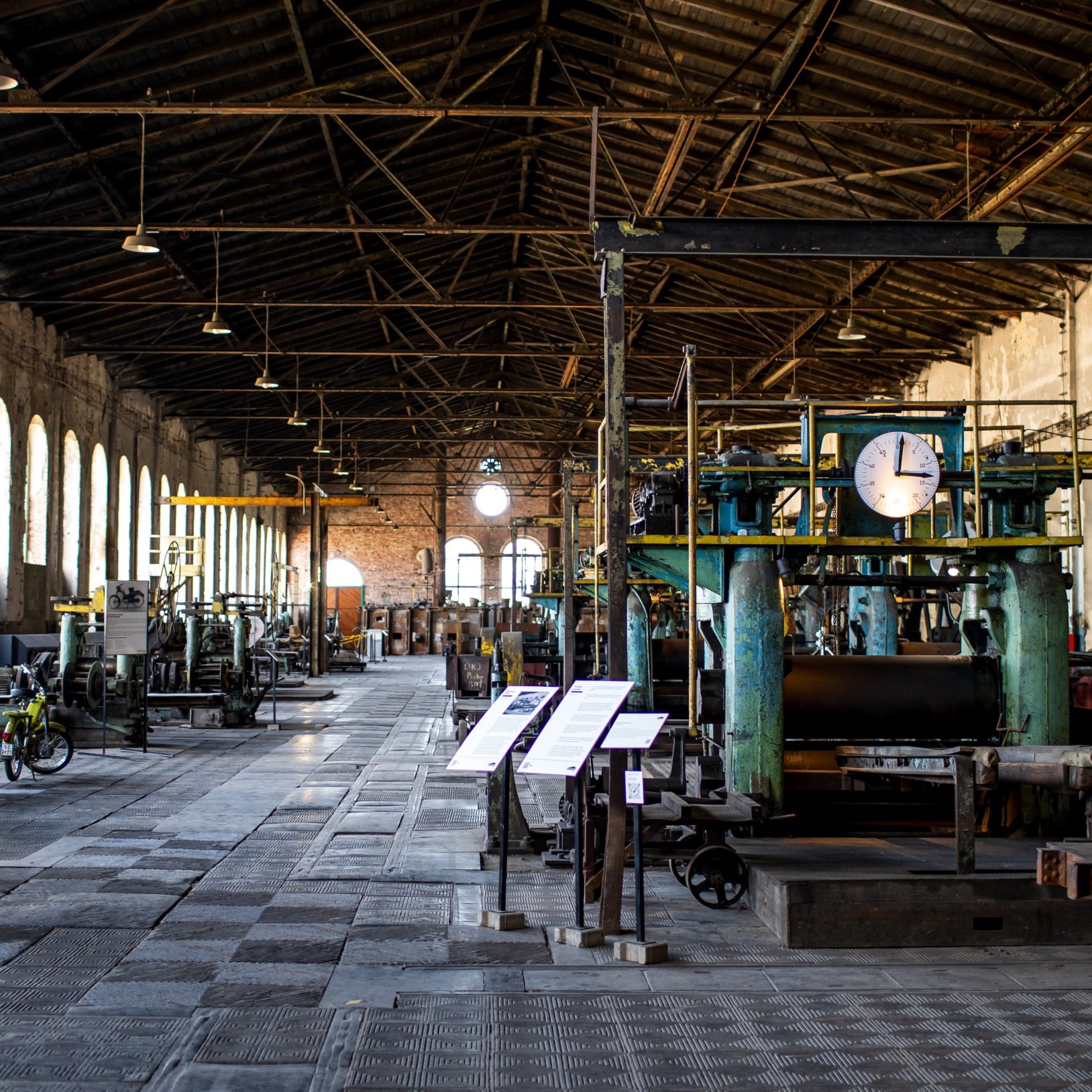 "Wnętrze dużej hali przemysłowej z wysokim, stalowym stropem i rzędem okien po lewej stronie; wzdłuż posadzki stoją zabytkowe maszyny i stanowiska z tablicami informacyjnymi, a na pierwszym planie po prawej widać dużą zieloną maszynę z okrągłym zegarem."
