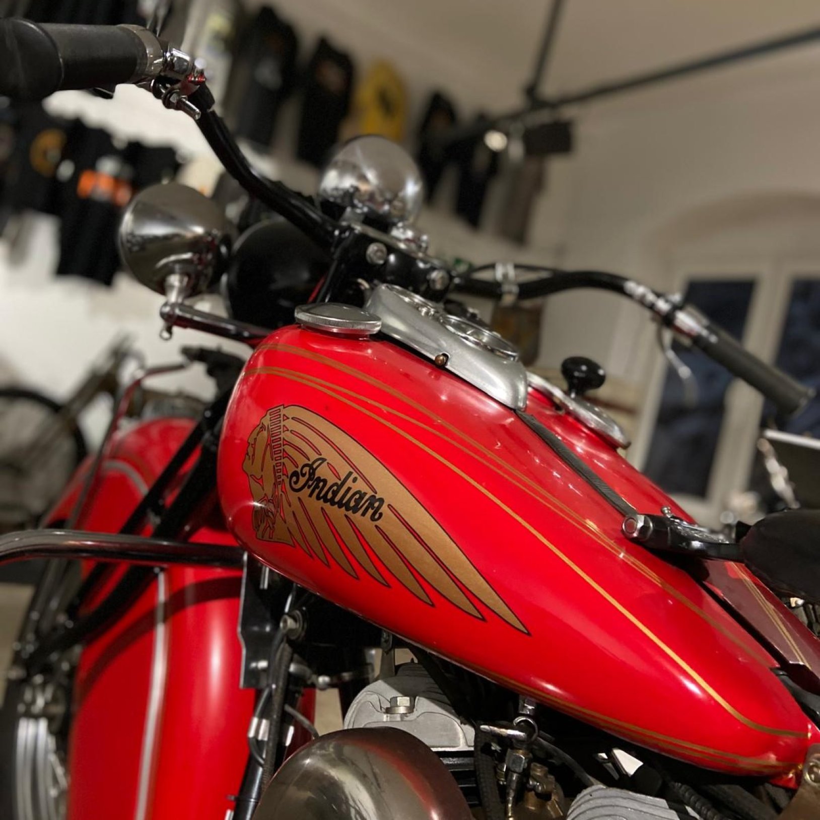 "Zbliżenie na czerwony, lakierowany zbiornik paliwa motocykla z logo „Indian” i złotymi liniami; widać także chromowane elementy kierownicy i reflektora, a tło jest rozmyte jak w pomieszczeniu wystawowym."