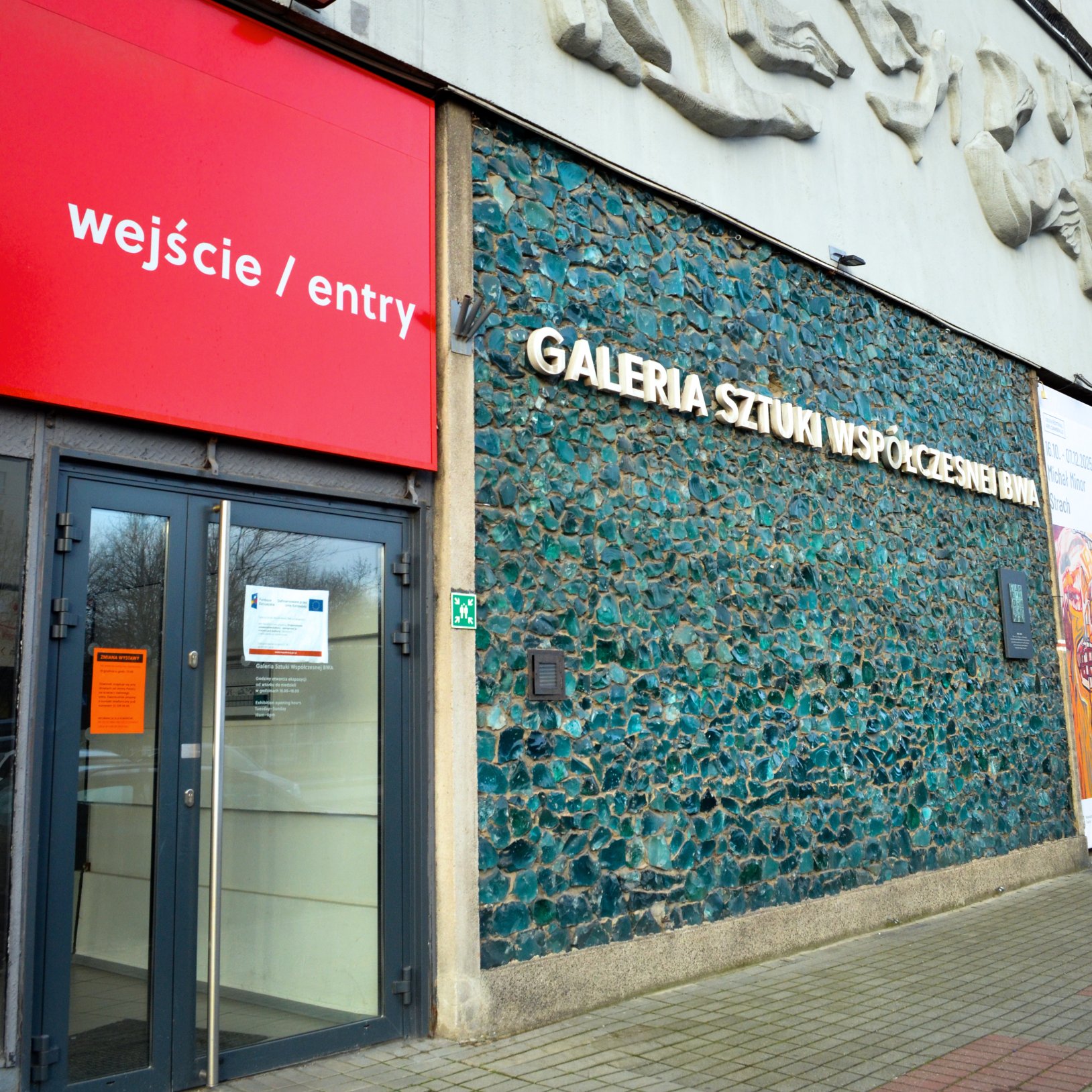 "Wejście do Galerii Sztuki Współczesnej z czerwonym szyldem 'wejście / entry' nad drzwiami. Po prawej stronie znajduje się ściana wyłożona zielonkawo-niebieskimi kawałkami szkła oraz srebrny napis 'GALERIA SZTUKI WSPÓŁCZESNEJ'."