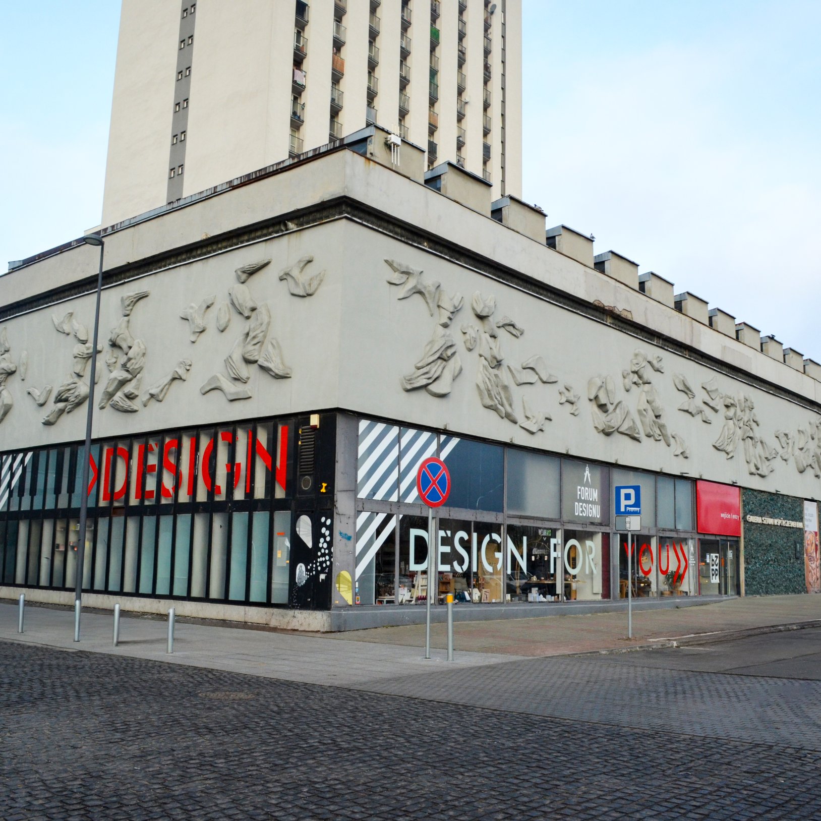 "Budynek z wysoką wieżą i parterową częścią z dużymi witrynami, w których widoczne są napisy 'DESIGN FOR' oraz 'FORUM DESIGNU'. Fasada ozdobiona jest płaskorzeźbami przedstawiającymi postacie w ruchu.