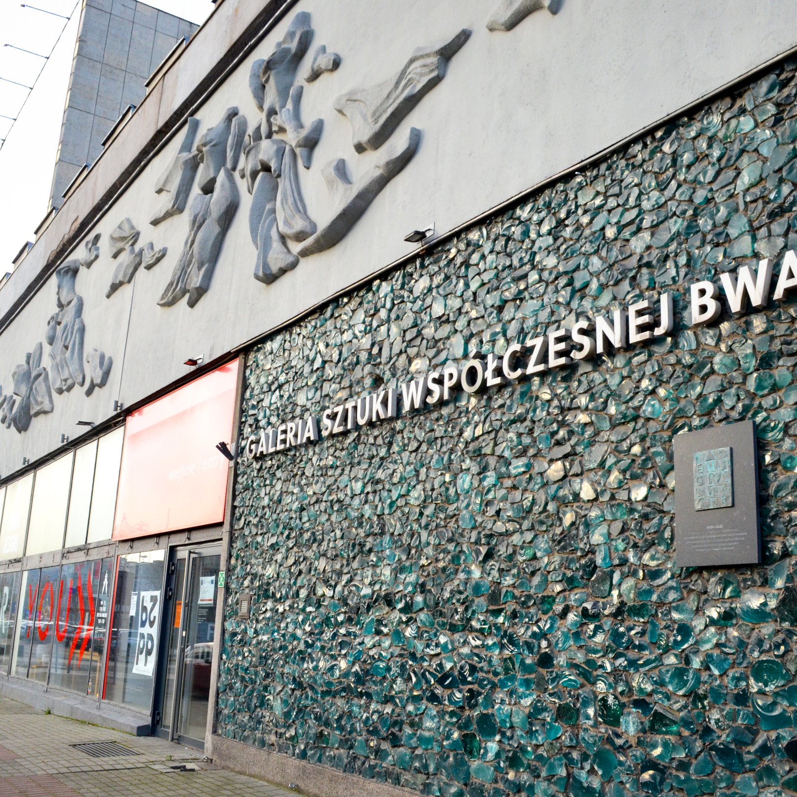 "Fragment elewacji budynku z dużym napisem „GALERIA SZTUKI WSPÓŁCZESNEJ BWA” na ścianie wyłożonej zielonkawą mozaiką z nieregularnych kawałków; powyżej widoczne szare płaskorzeźby, a po lewej szklane wejście i witryny przy chodniku."