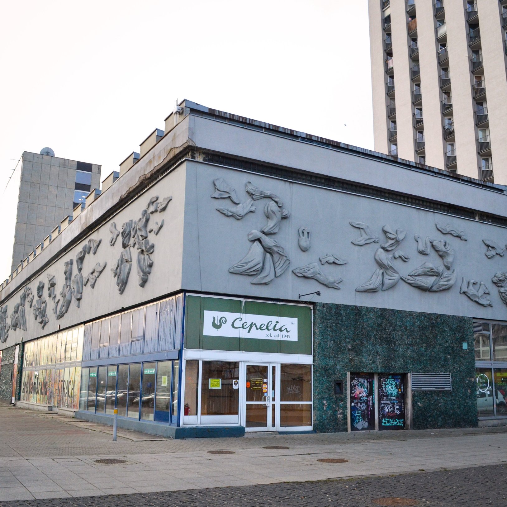 "Widok na narożny, niski budynek z szarymi płaskorzeźbami na górnej części elewacji i zielonkawą mozaiką na parterze; na froncie wejście z szyldem „Cepelia, rok zał. 1949”, obok drzwi z graffiti, w tle wysoki blok mieszkalny."