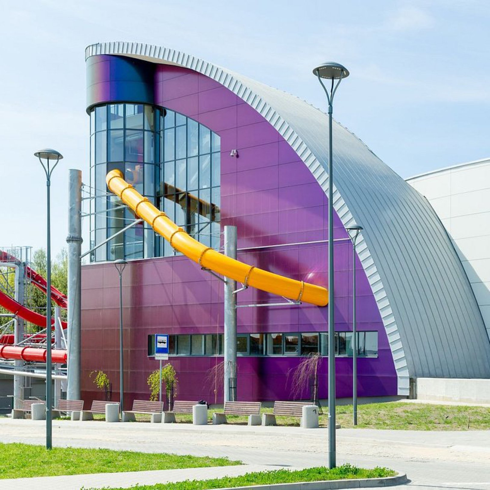 Nowoczesna fasada Parku Wodnego Tychy z wygiętym dachem i fioletowo-srebrną elewacją. Po lewej stronie budynku widoczna jest długa, żółta zjeżdżalnia wodna.