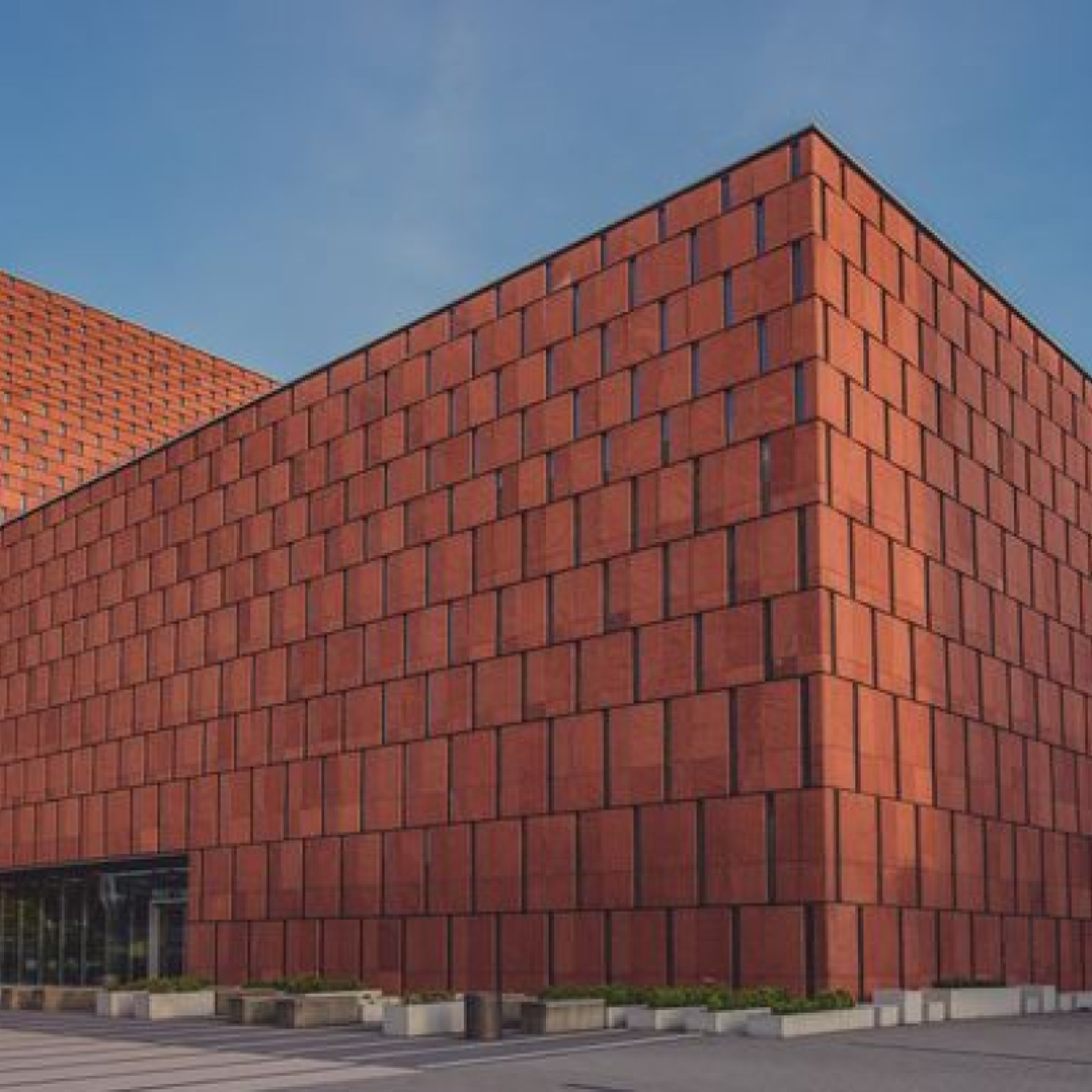 Zewnętrzna część nowoczesnego budynku biblioteki pokryta czerwonymi płytkami elewacyjnymi. Bryła budynku ma kanciaste, geometryczne kształty. Na pierwszym planie widoczny chodnik i kulista betonowa rzeźba.