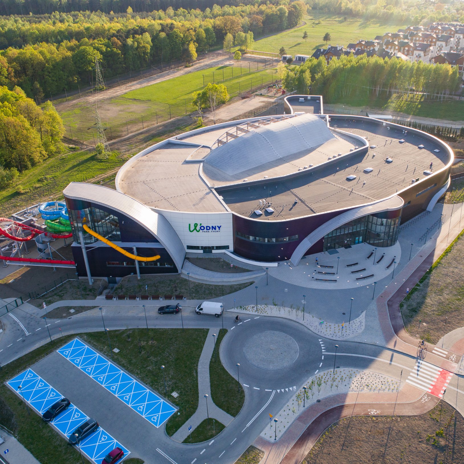 Widok z lotu ptaka na nowoczesny kompleks basenowy Parku Wodnego Tychy. Budynek ma opływowy kształt, a na zewnątrz widoczne są kolorowe zjeżdżalnie. Otoczony jest drogami, parkingiem i terenami zielonymi.