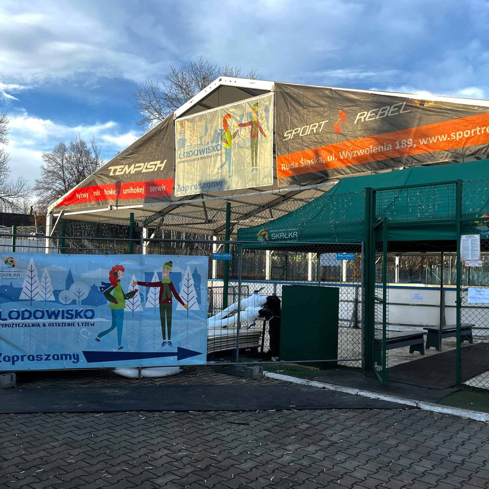Wejście na odkryte lodowisko ogrodzone siatką, z zieloną bramką i zadaszeniem; na hali nad lodowiskiem wiszą banery reklamowe, a na ogrodzeniu wisi plakat Lodowisko z rysunkami łyżwiarzy i strzałką kierunku.