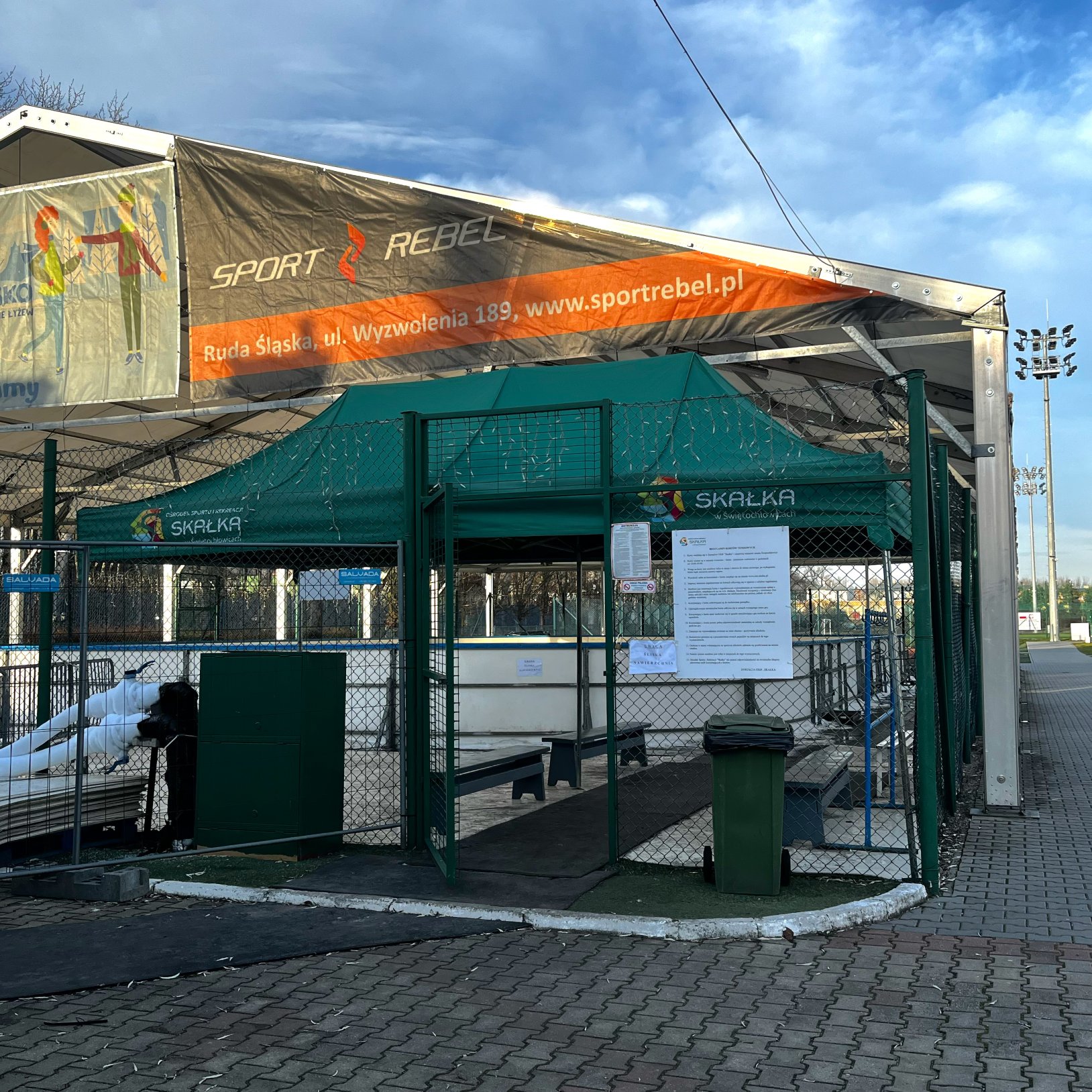 Widok na bramę wejściową na lodowisko z zielonym namiotem i tablicą z regulaminem na ogrodzeniu; nad konstrukcją zadaszenia widać duży baner SPORT REBEL, a przed wejściem stoi zielony kosz na śmieci.