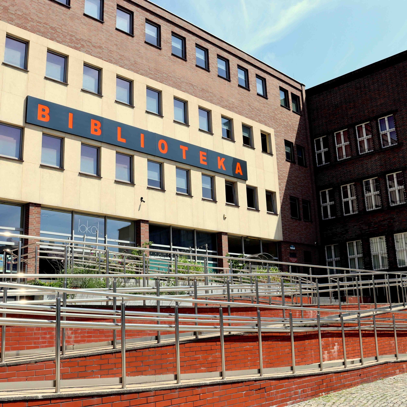 "Nowoczesny budynek biblioteki z fasadą z beżowych i ceglastych paneli. Na elewacji widoczny duży napis 'BIBLIOTEKA' z pomarańczowymi literami. Przed budynkiem znajdują się metalowe barierki i schody."