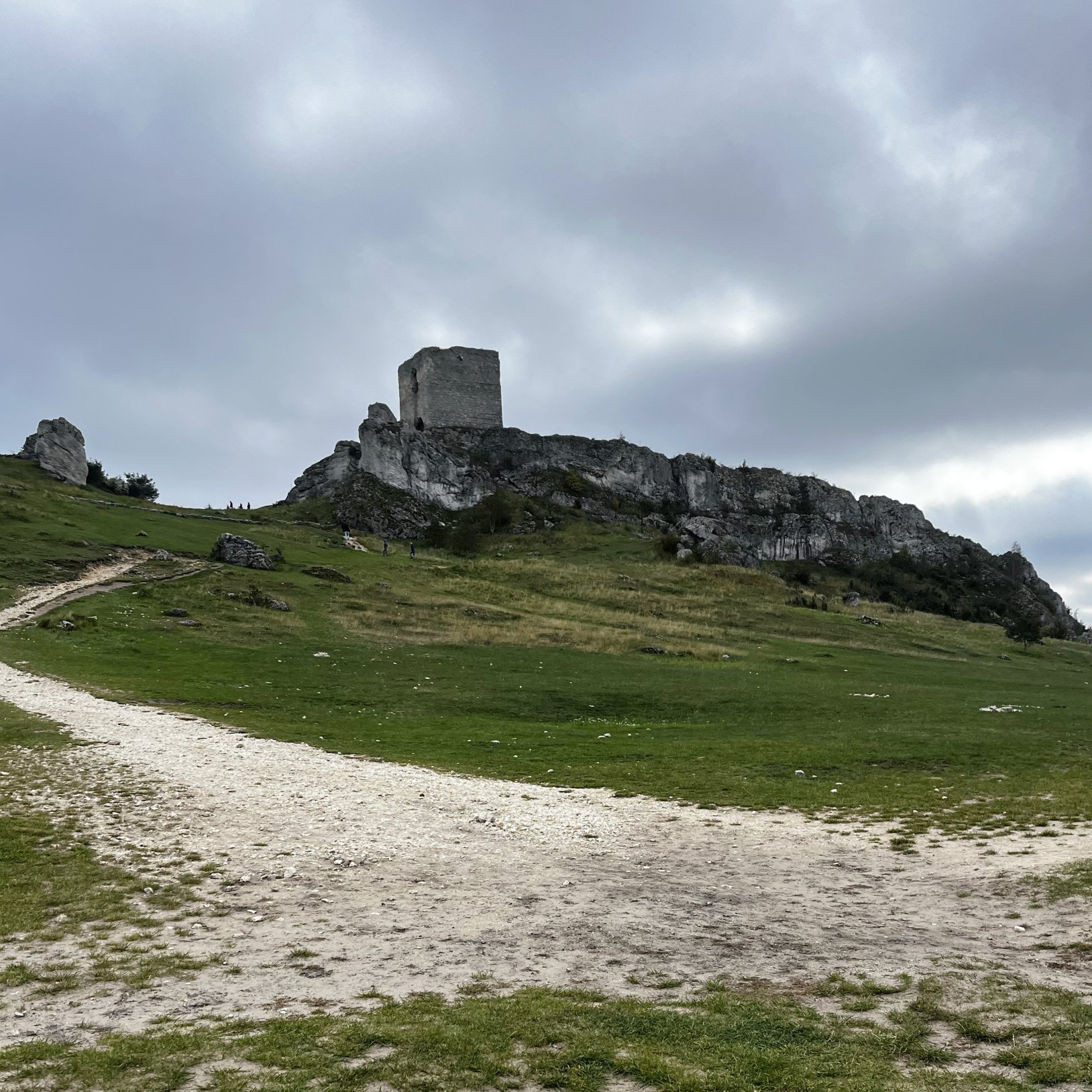 "Ruiny kamiennej wieży na skalnym grzbiecie widziane z oddali; przez zielone zbocze biegnie jasna, piaszczysta ścieżka, a w tle widać ciężkie, szare chmury."   }