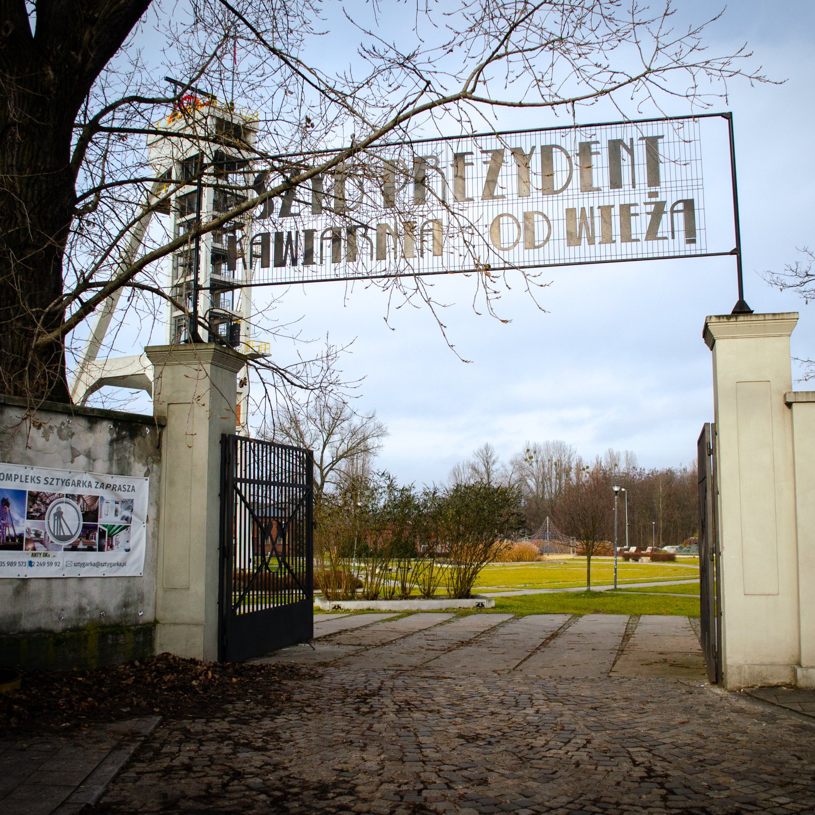 "Wejście przez otwartą bramę między jasnymi słupami, nad którą wisi metalowy napis „SZTYGARKA KAWIARNIA POD WIEŻĄ”; w tle widać alejki i zieleń, a po lewej częściowo widoczna jest wieża szybowa."