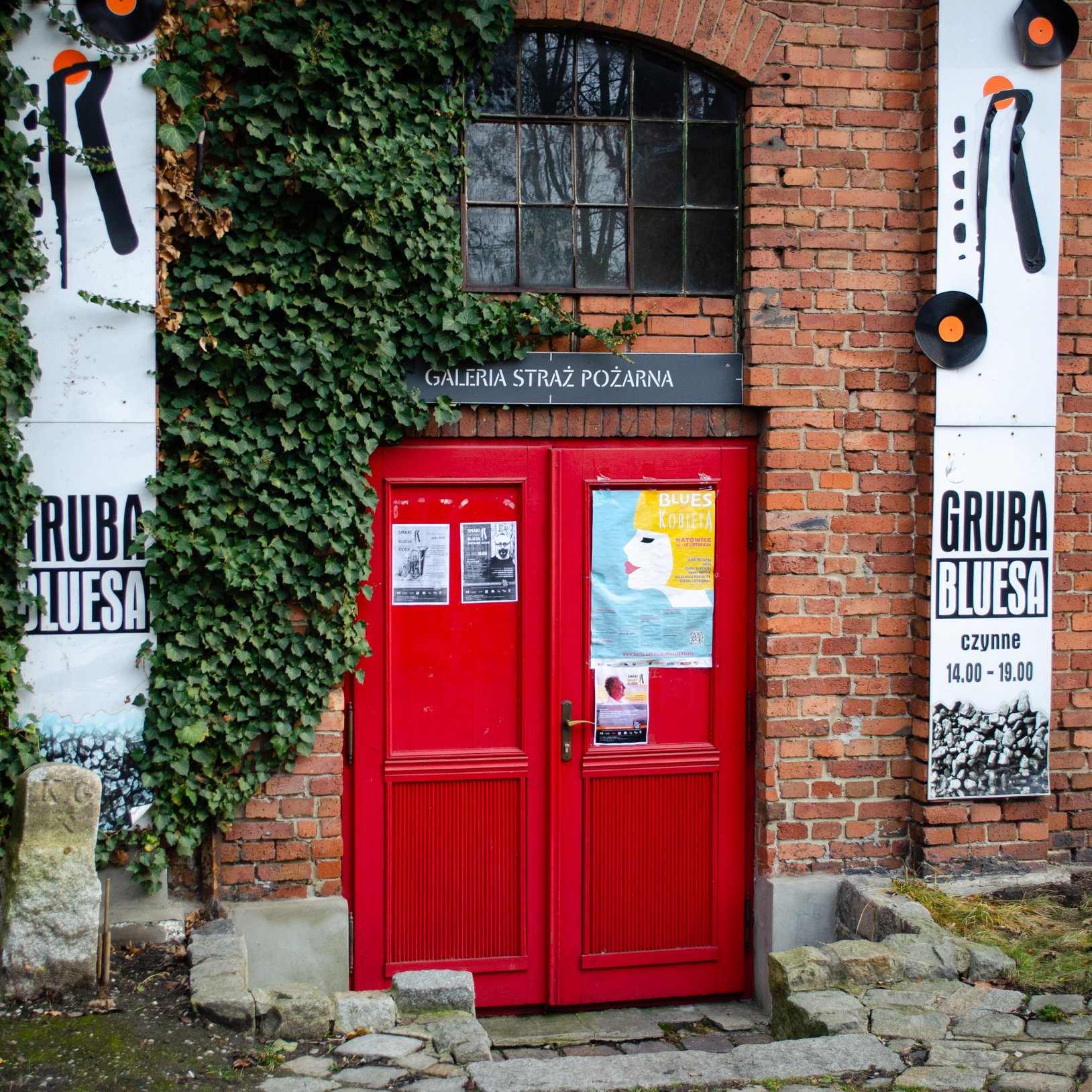 "Zbliżenie na czerwone drzwi w ceglanej elewacji z tabliczką „GALERIA STRAŻ POŻARNA”; po lewej bluszcz na ścianie, a po prawej pionowy szyld „GRUBA BLUESA” z informacją o godzinach otwarcia."