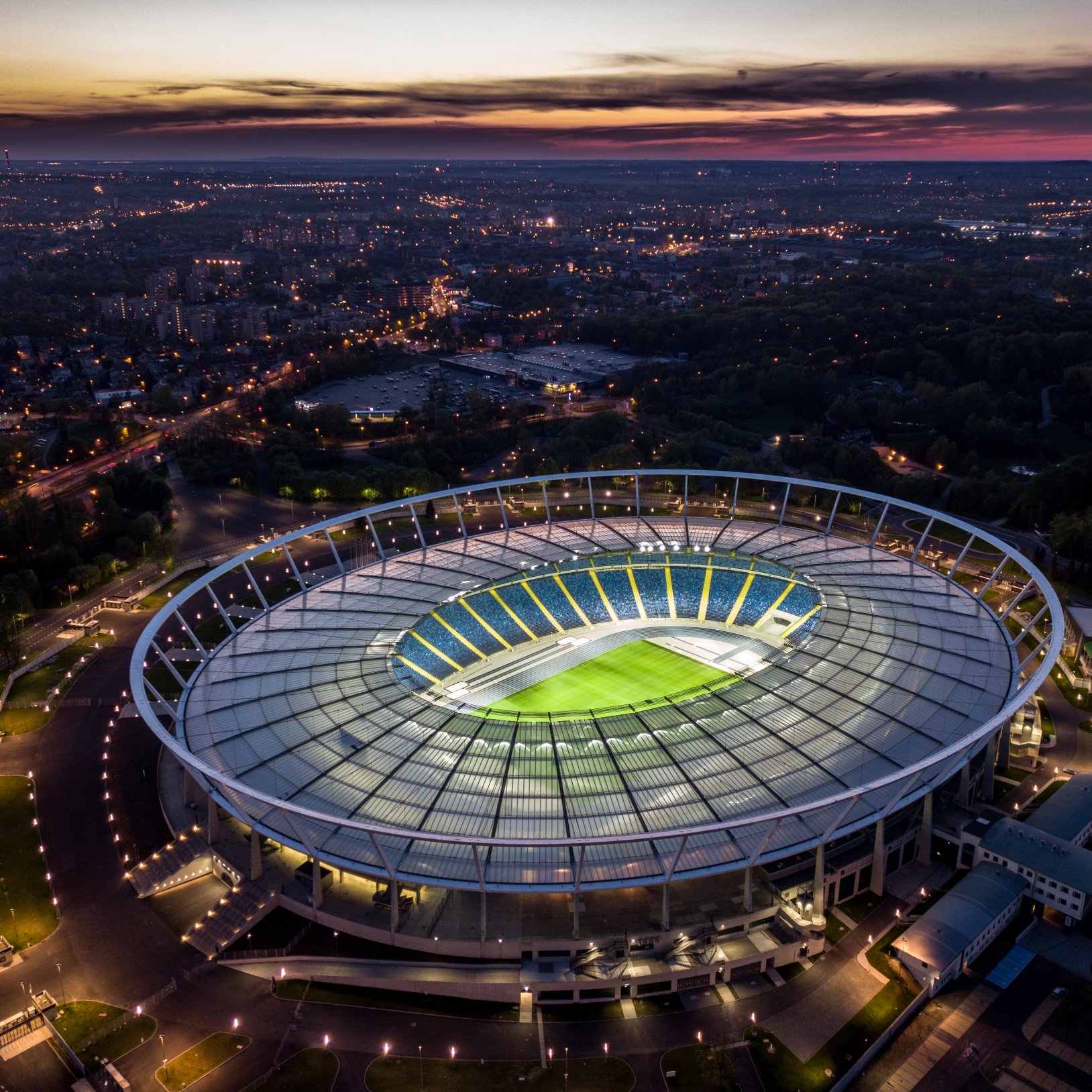 "Nocna panorama Stadionu Śląskiego z lotu ptaka. Stadion oświetlony wewnątrz, otoczony ciemną zabudową miasta i rozświetlonymi ulicami. Widoczna eliptyczna konstrukcja dachu i układ trybun."