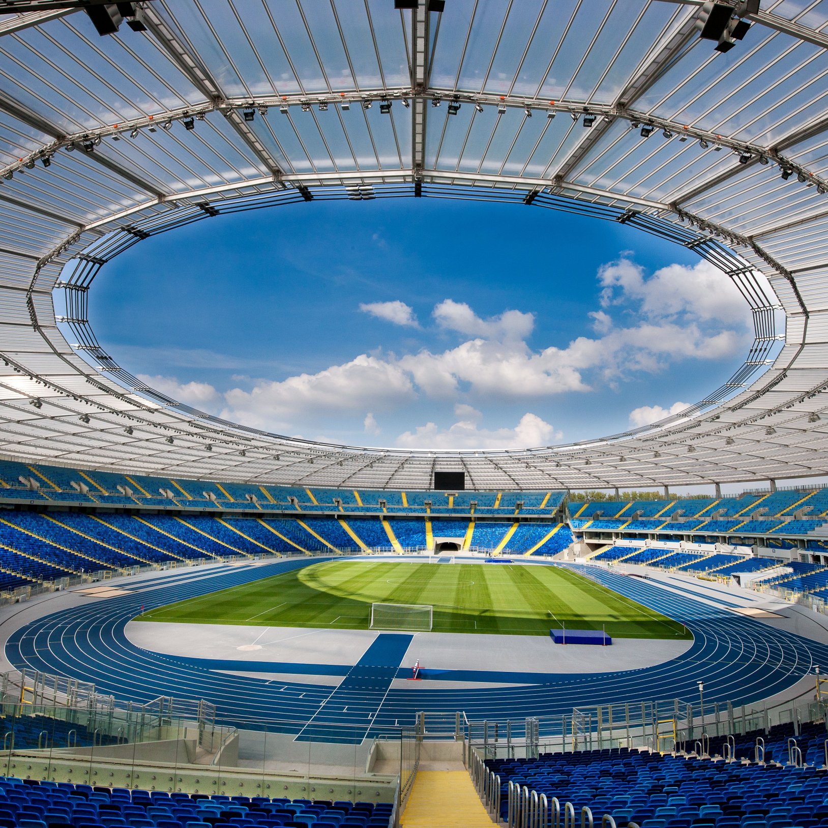 "Widok z poziomu murawy Stadionu Śląskiego na trybuny i dach. Na pierwszym planie trawiasta nawierzchnia boiska z białą linią, w tle błękitne niebo z chmurami."