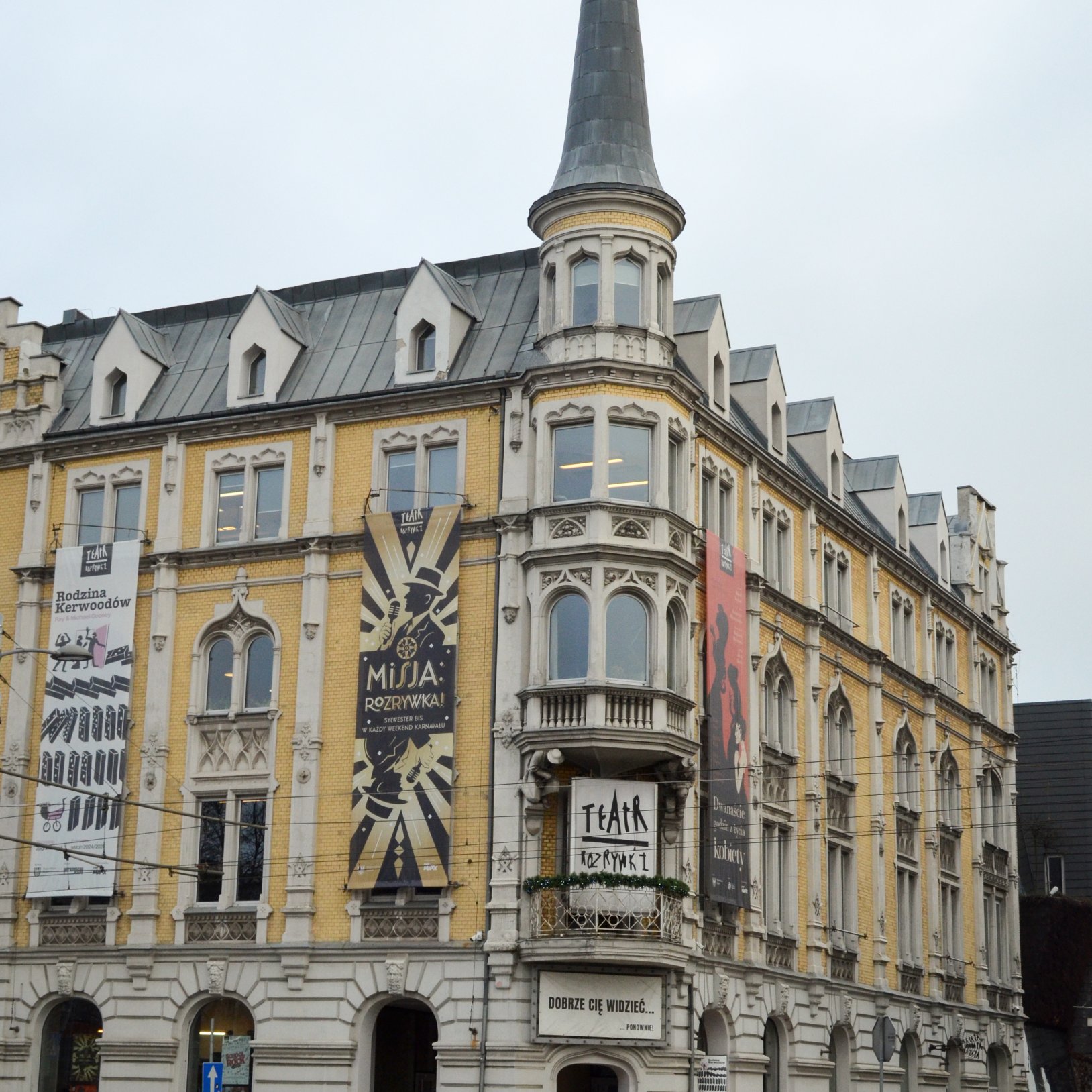 "Zbliżony widok na narożnik Teatru Rozrywki z ozdobnymi oknami i wieżyczką; na elewacji widoczne są pionowe banery reklamujące spektakle oraz szyld „TEATR ROZRYWKI”."