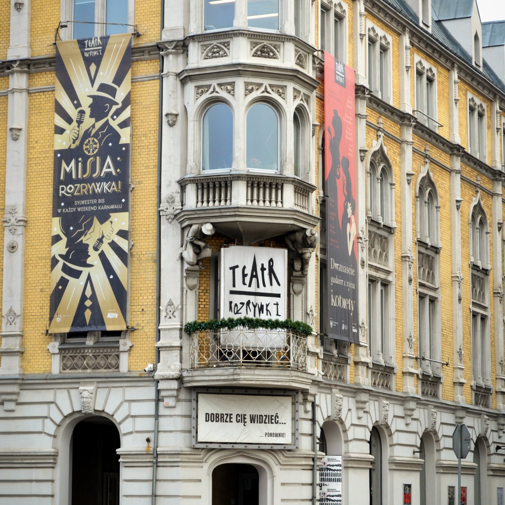 "Fragment fasady Teatru Rozrywki z wykuszem i balkonem; obok wiszą duże banery, w tym plakat z napisem „MISJA: ROZRYWKA!”, a pod balkonem znajduje się tablica z tekstem „DOBRZE CIĘ WIDZIEĆ…”."