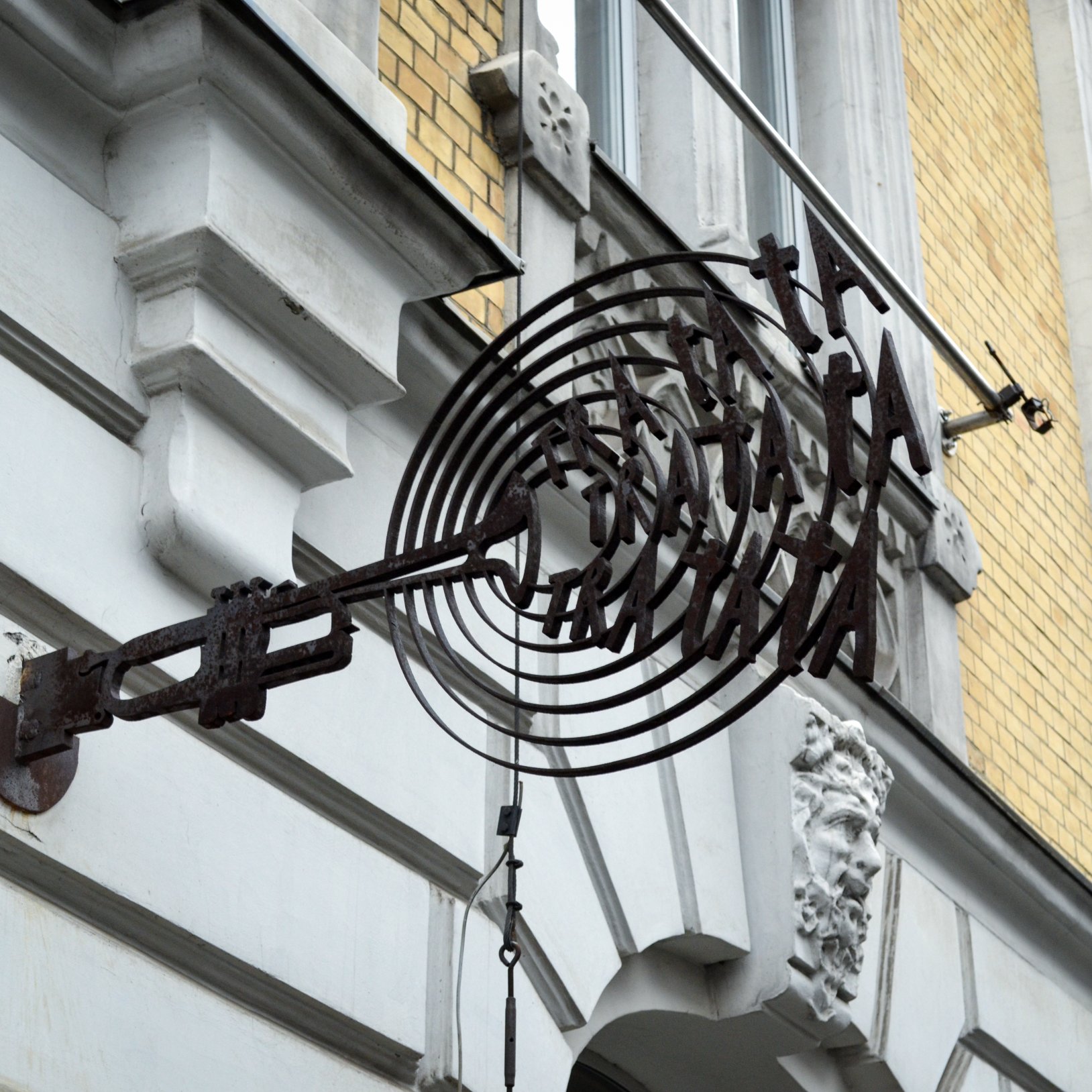 "Metalowy, ozdobny szyld w kształcie klucza z napisem „TEATR ROZRYWKI” zamocowany do elewacji; w tle widoczna jest jasna ściana budynku i dekoracyjna rzeźbiona głowa na fasadzie."