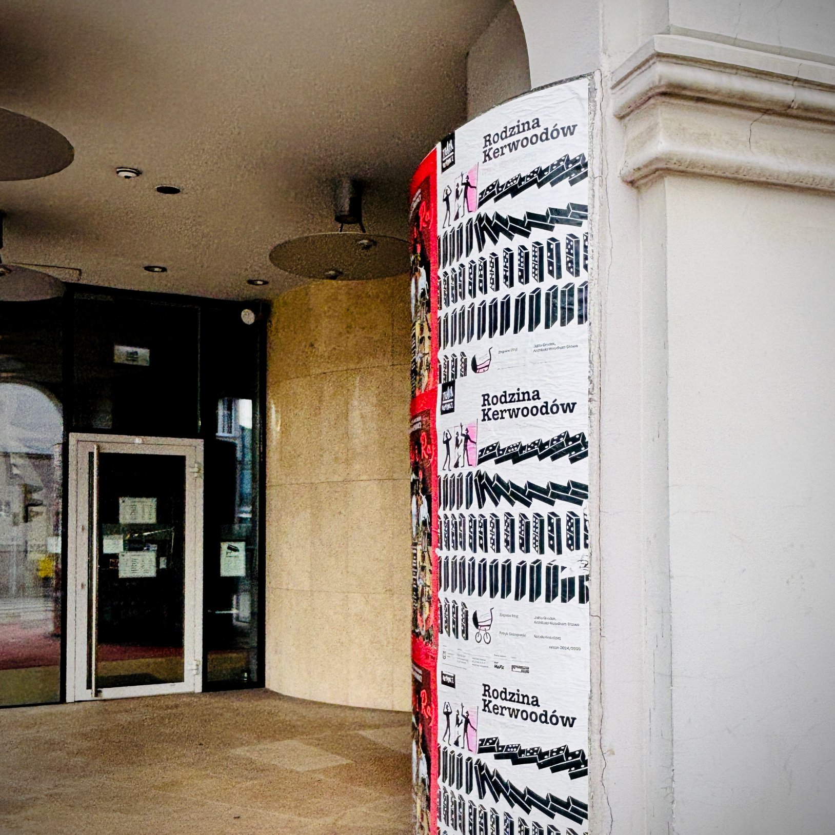 "Wejście do budynku pod arkadą; na filarze naklejone plakaty z tytułem „Rodzina Kerwoodów”, a obok widać przeszklone drzwi i oświetlenie sufitowe w podcieniu."