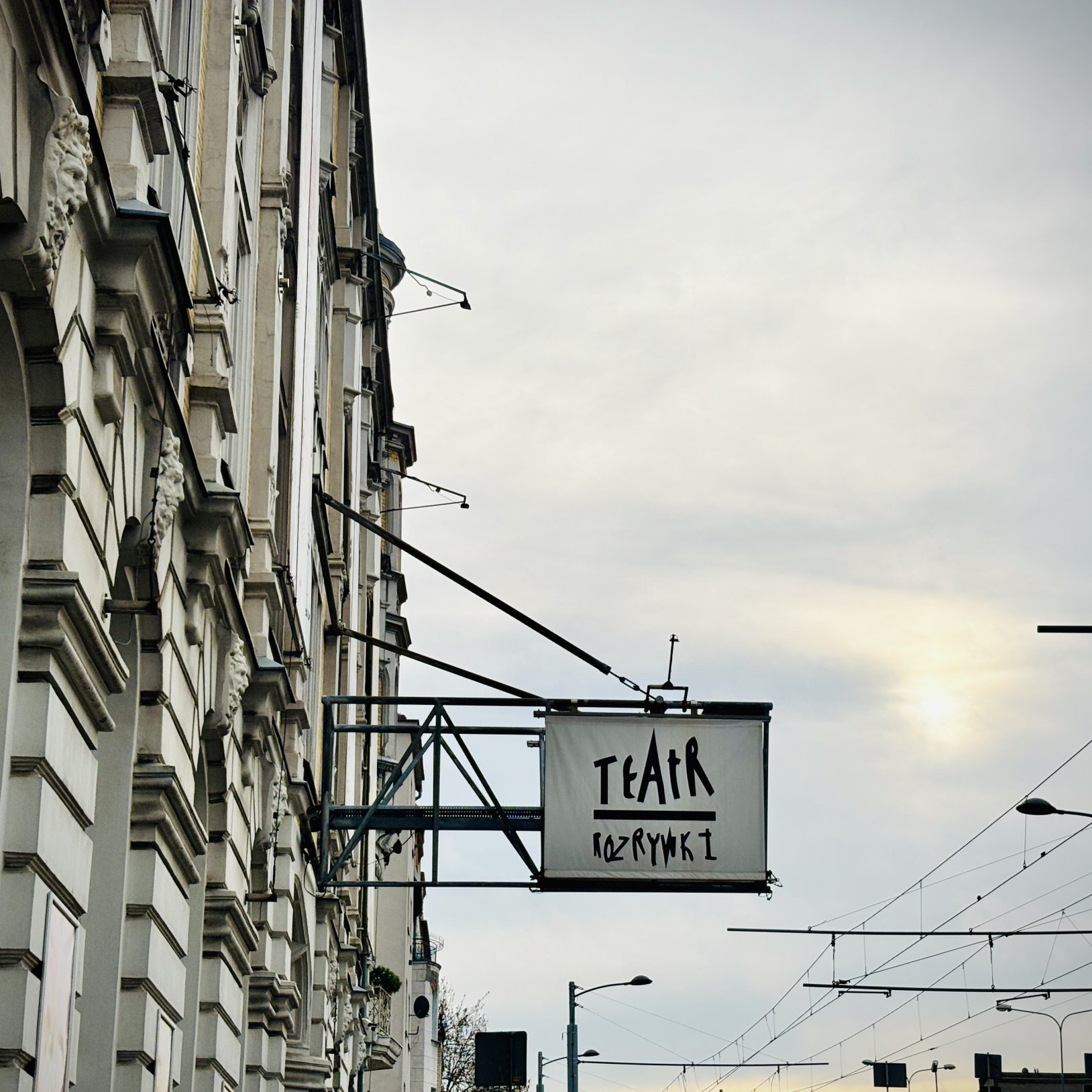 "Boczny widok na elewację kamienicy z wysięgnikiem, na którym wisi kwadratowy szyld „TEATR ROZRYWKI”; w tle widać przewody trakcyjne i latarnie uliczne pod pochmurnym niebem."
