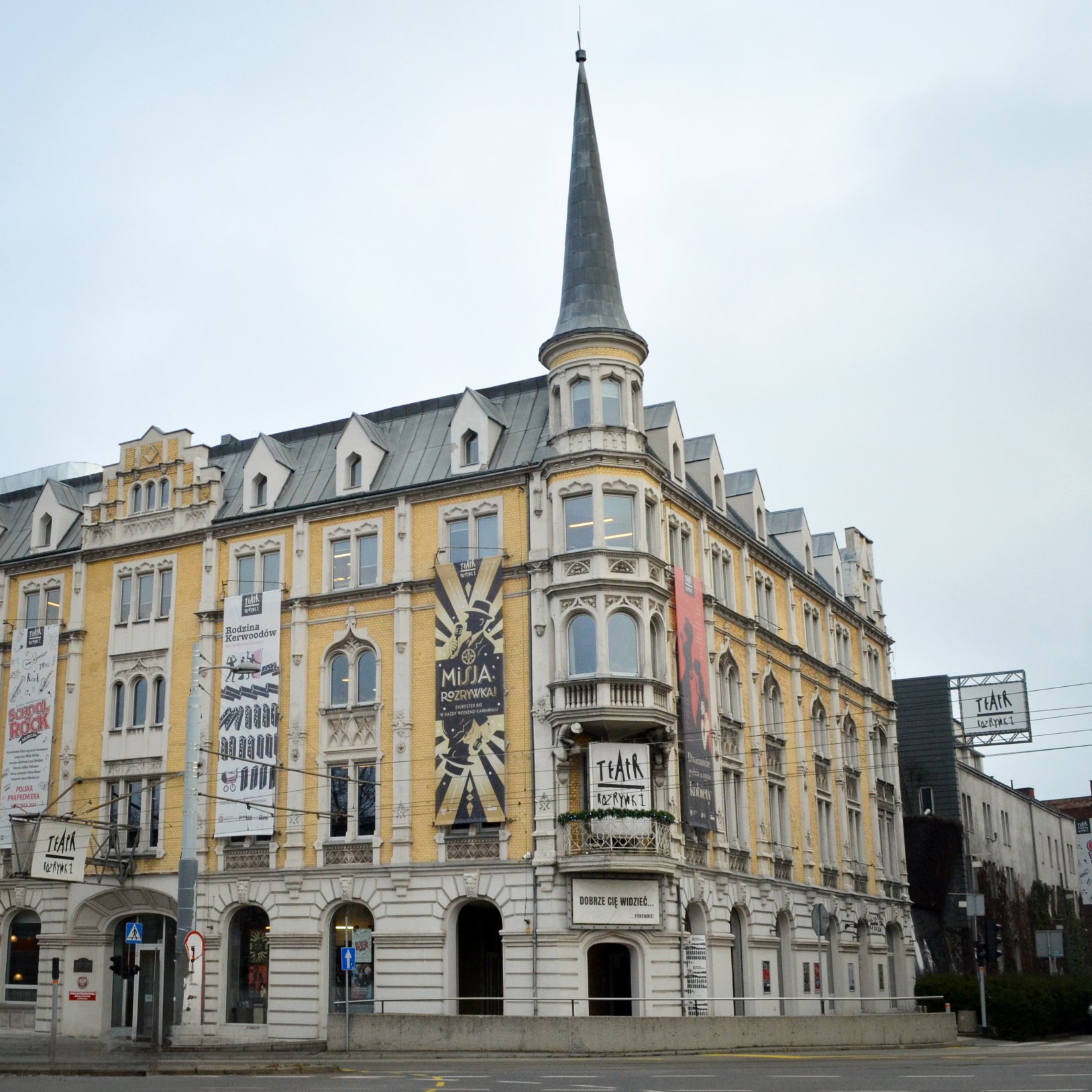 "Szeroki widok na zabytkowy, żółto-biały budynek z narożną wieżyczką i wysoką iglicą; na fasadzie wiszą duże plakaty oraz szyld „TEATR ROZRYWKI”, a przed budynkiem widać ulicę i przewody trakcyjne pod jasnym niebem."