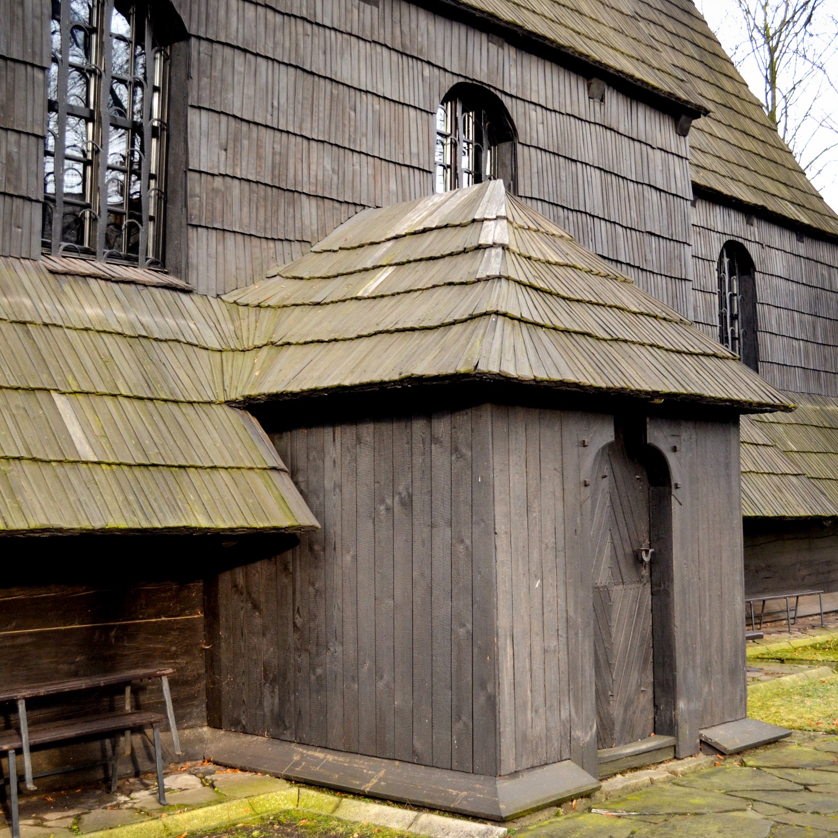 "Zbliżenie na boczne wejście drewnianego kościoła: mały ganek z gontowym daszkiem i drewnianymi drzwiami; obok widać ławkę oraz fragment ściany z oknami w łukowych wnękach."