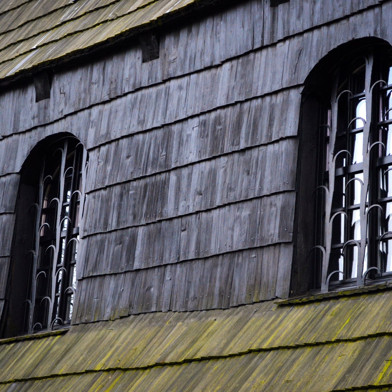 "Detal elewacji drewnianego kościoła z dwoma wysokimi, łukowymi oknami z metalowymi kratami; na pierwszym planie widoczny gontowy daszek z zielonym nalotem."