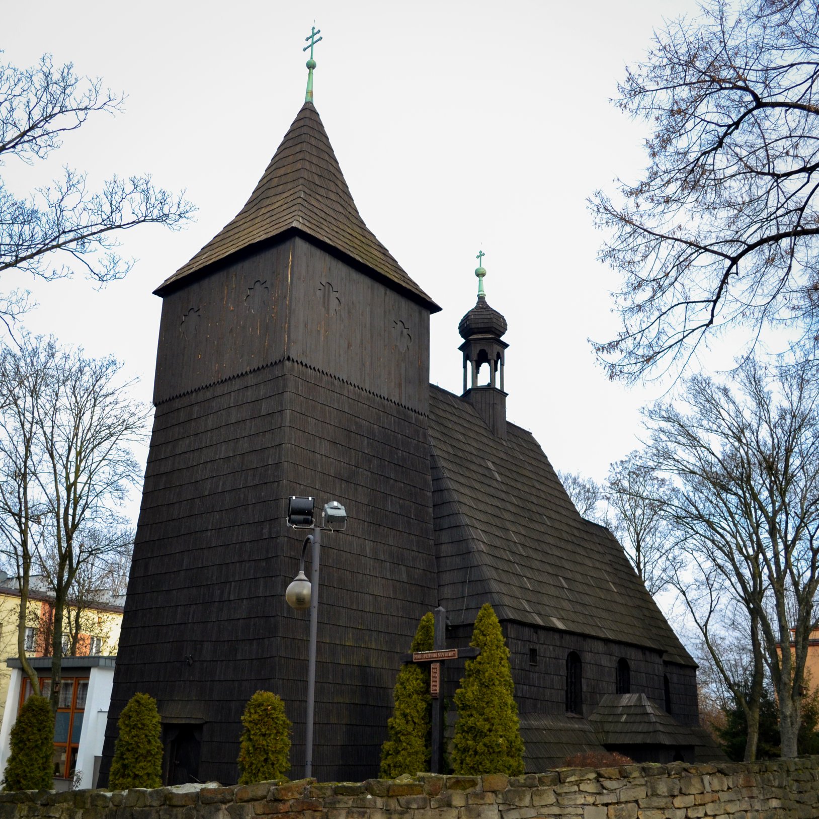 "Widok z przodu na drewniany kościół z wysoką wieżą i sygnaturką, oba zwieńczone krzyżami; przed budynkiem stoi niski kamienny mur i rząd iglaków, a w tle widać bezlistne drzewa."