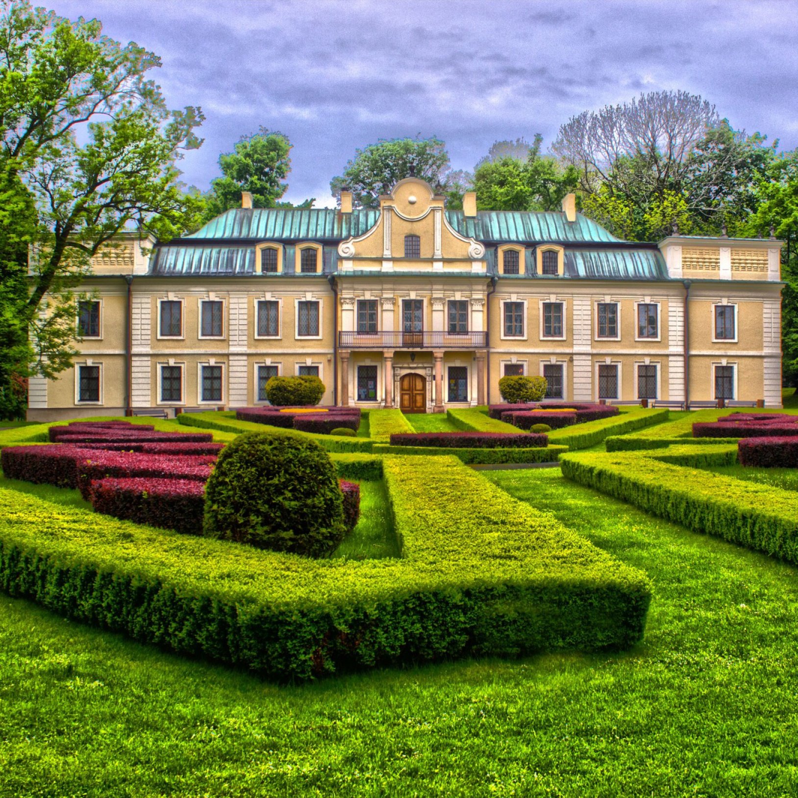 Widok na klasycystyczny pałac z jasną elewacją i zielonym dachem mansardowym, otoczony symetrycznym, zadbanym ogrodem w stylu francuskim. Przed pałacem znajdują się przycięte żywopłoty i krzewy tworzące geometryczne wzory.