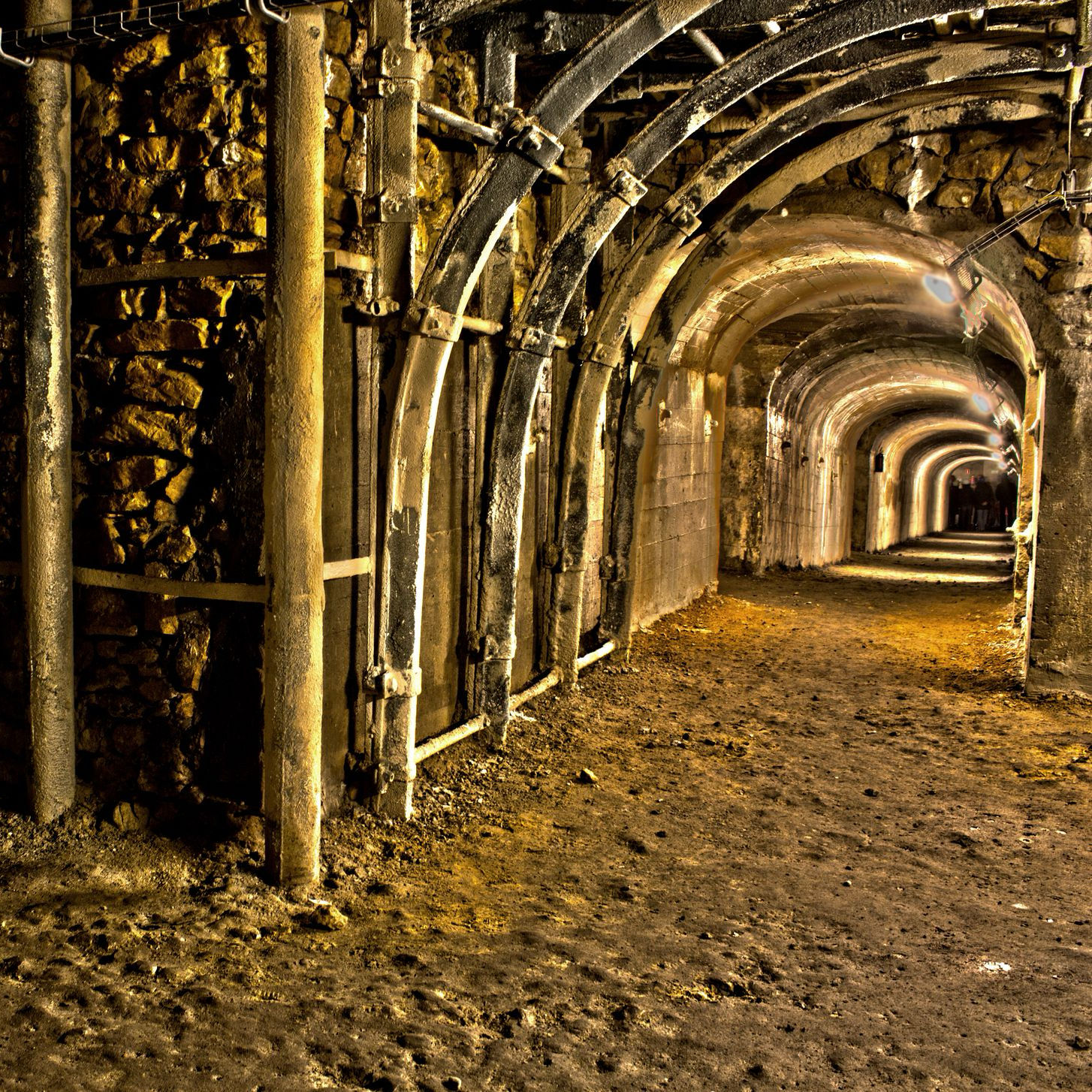 Podziemny tunel z kamiennymi i betonowymi ścianami oraz łukowatym sklepieniem. Korytarz oświetlony jest sztucznym światłem zamontowanym wzdłuż sufitu. Konstrukcja wsparta metalowymi belkami i rurami. Podłoże nierówne, pokryte ziemią i żwirem.