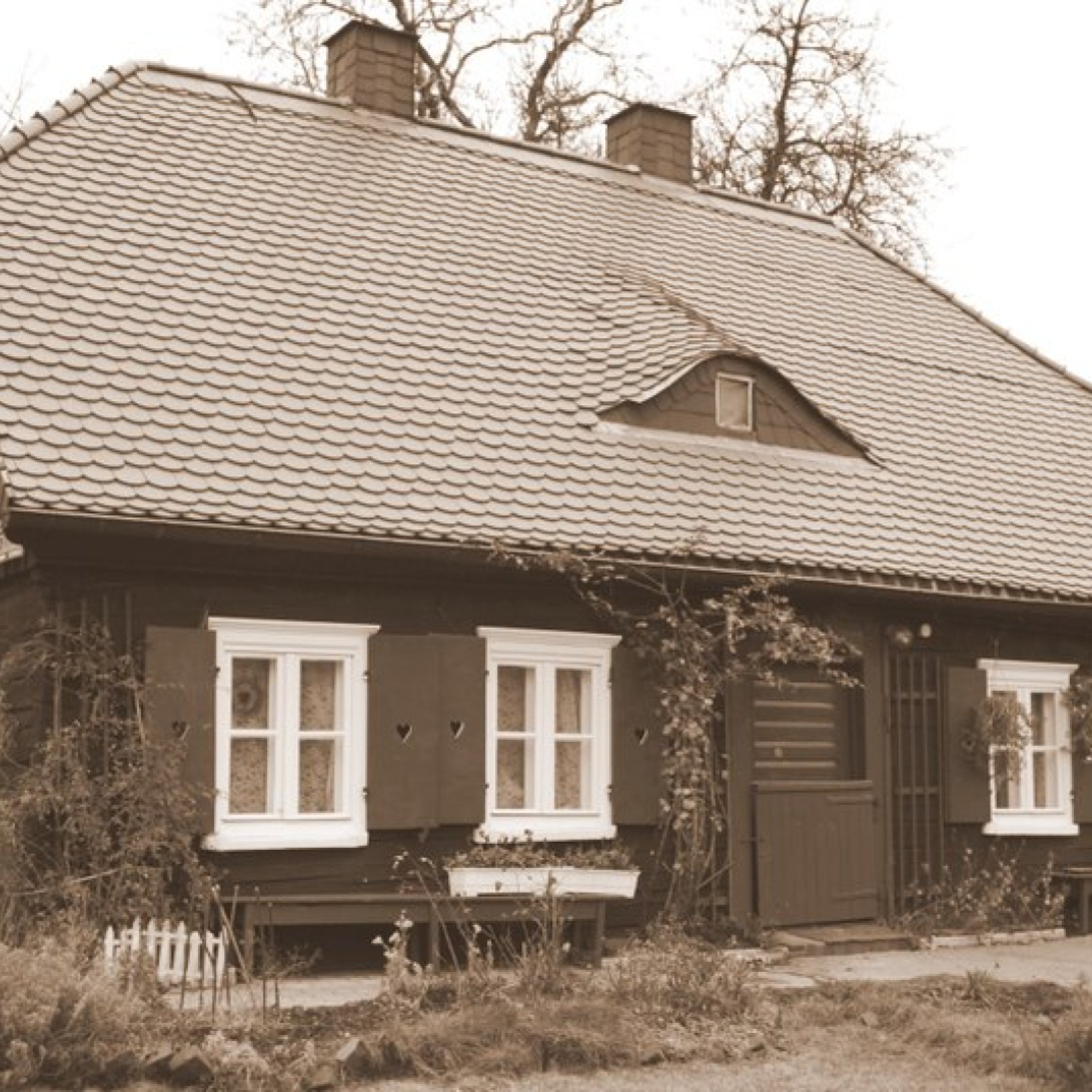fotografia w sepii przedstawiający drewniany dom Matki Ewy, dom ze spadzistym dachem z wolim okiem i drewnianymi okiennicami, wokół domu roślinność