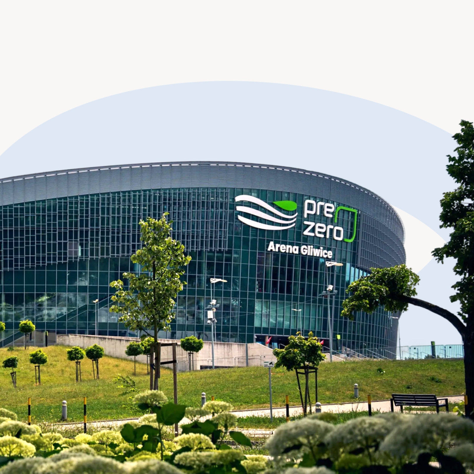 "Zdjęcie przedstawia zewnętrzną fasadę PreZero Areny Gliwice – nowoczesnego budynku o zaokrąglonym kształcie z przeszkloną elewacją. Przed areną widoczna jest zieleń: drzewa, krzewy i trawnik. Na fasadzie umieszczone jest logo z nazwą obiektu."