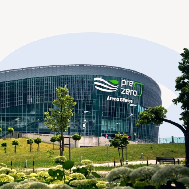 "Zdjęcie przedstawia zewnętrzną fasadę PreZero Areny Gliwice – nowoczesnego budynku o zaokrąglonym kształcie z przeszkloną elewacją. Przed areną widoczna jest zieleń: drzewa, krzewy i trawnik. Na fasadzie umieszczone jest logo z nazwą obiektu."