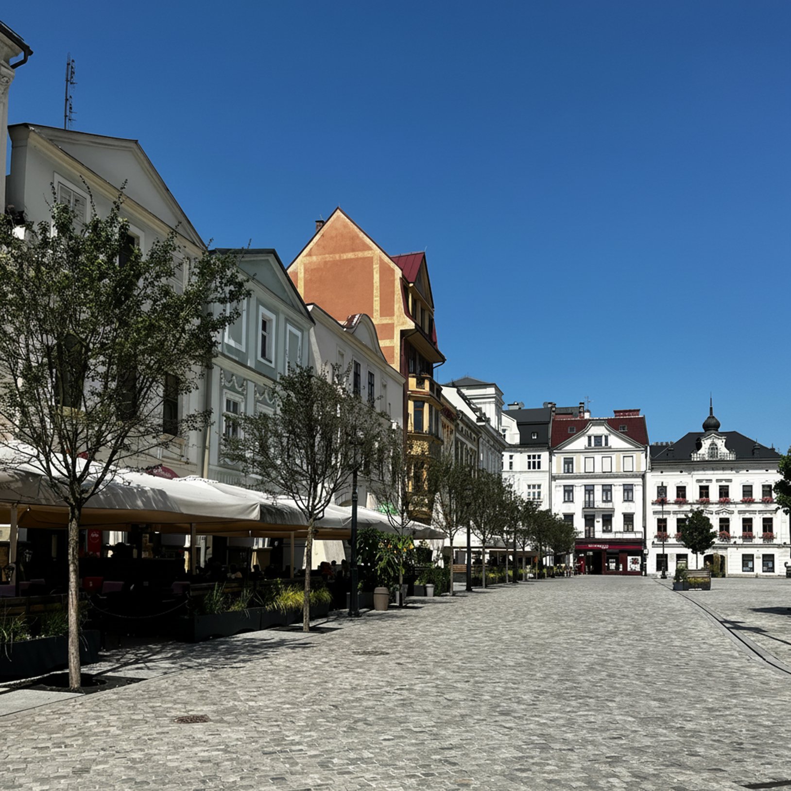 "Perspektywa wzdłuż brukowanego rynku z rzędem kamienic po lewej i ogródkami kawiarnianymi pod jasnymi parasolami; w głębi widoczne białe, ozdobne fasady budynków, a całość oświetla słońce przy czystym, niebieskim niebie."