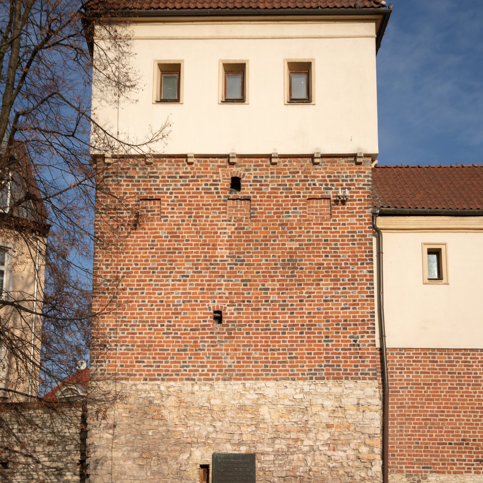 "Zbliżenie na jedną z wież budynku wykonaną z czerwonej cegły i kamienia, z jasną górną częścią i trzema oknami. Na dole widoczna tablica pamiątkowa zamontowana na ścianie oraz osoba siedząca na ławce u podnóża wieży. Obok drzewo bez liści i fragment sąsi