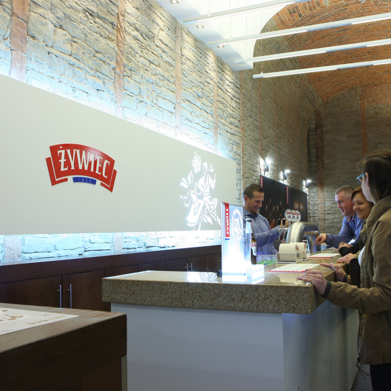 "Nowoczesna lada degustacyjna z logo Żywiec wewnątrz Muzeum Browaru Żywiec. Przy barze stoi kilka osób degustujących piwo, obsługiwanych przez pracownika. Wnętrze wykończone kamiennymi ścianami i ceglanym sklepieniem łukowym."