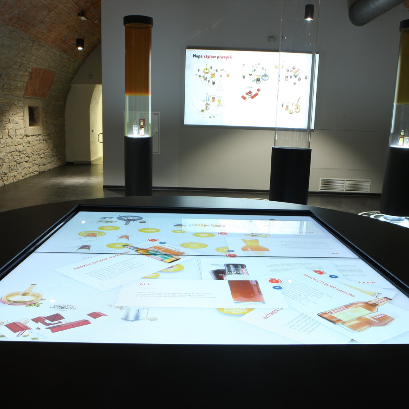 "Interaktywna sala wystawiennicza w Muzeum Browaru Żywiec. Na pierwszym planie znajduje się duży dotykowy stół multimedialny prezentujący informacje o stylach piwa. W tle widoczne są wysokie tuby z eksponatami oraz plansze edukacyjne na ścianie."   }