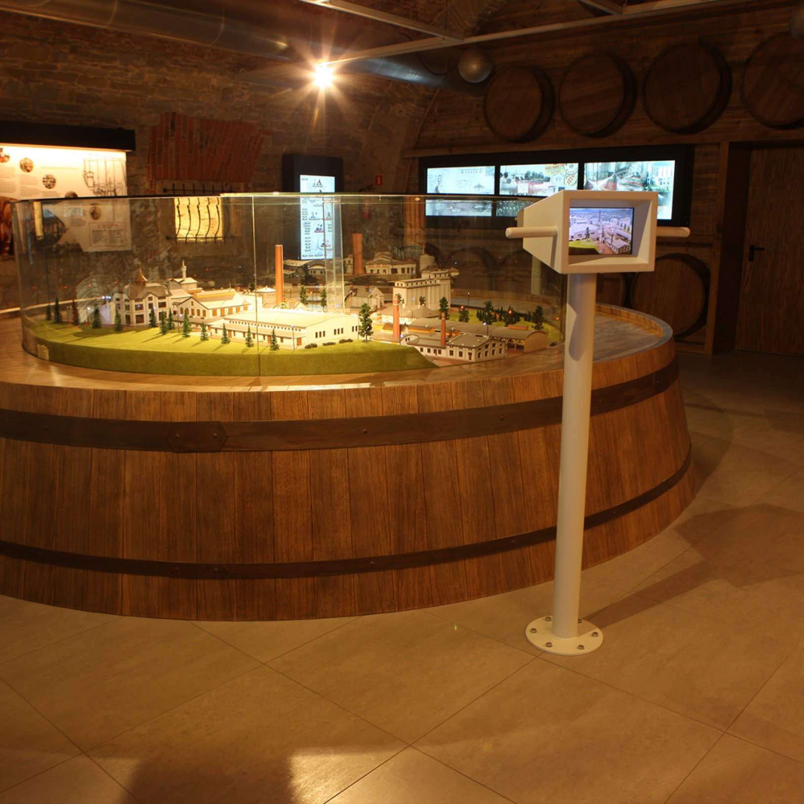 "Makieta dawnego browaru umieszczona w dużej, drewnianej konstrukcji przypominającej beczkę. Makieta przedstawia zabudowania i infrastrukturę zakładu. W tle znajdują się drewniane beczki na ścianie oraz ekrany i plansze informacyjne."