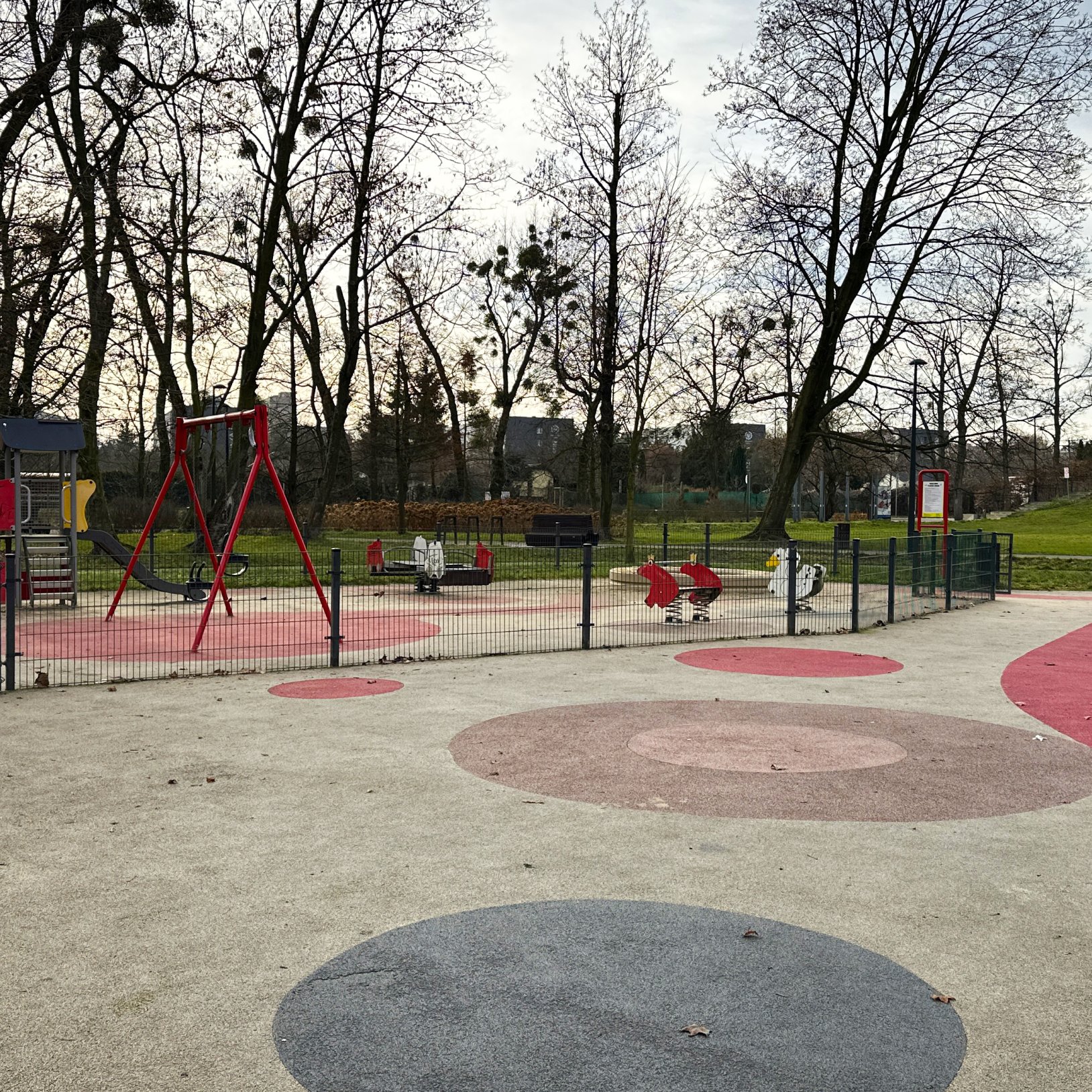 "Plac zabaw w Parku Chrobrego z kolorową, bezpieczną nawierzchnią i ogrodzonym terenem zabaw. W środku znajdują się huśtawki, bujaki sprężynowe oraz mała zjeżdżalnia. W tle widoczne ławki, trawniki i drzewa bez liści. Otoczenie sugeruje późną jesień lub w