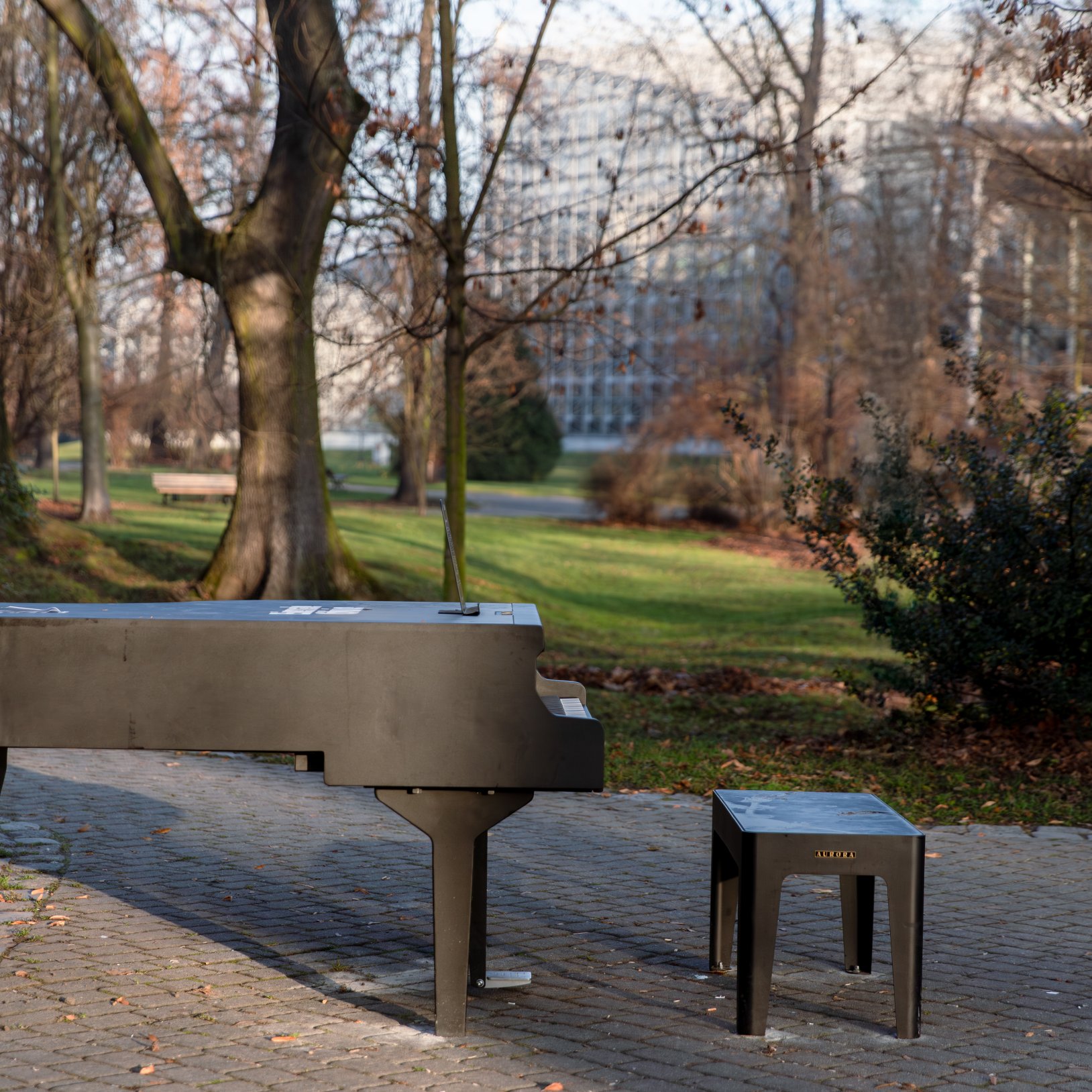 "Czarny fortepian i stołek ustawione na ścieżce w parku miejskim, w otoczeniu drzew i zieleni. W tle widoczny nowoczesny budynek biurowy. Teren parku pokryty jesiennymi liśćmi, a fortepian najprawdopodobniej jest instalacją artystyczną lub miejskim instru