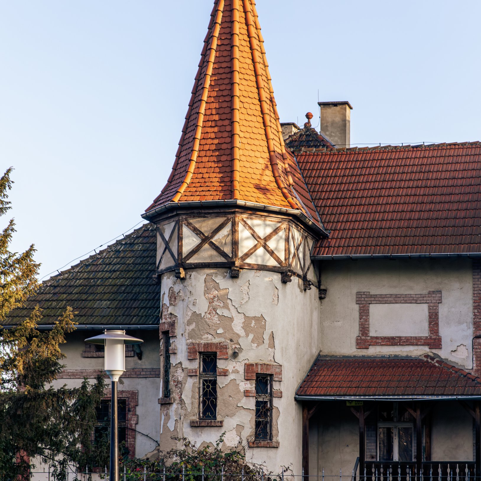 "Zabytkowy budynek z charakterystyczną wieżyczką pokrytą pomarańczową dachówką, zwieńczoną iglicą. Ściany wieży są zniszczone i popękane, z widocznymi śladami zużycia. Okna zdobione kratami, a wokół budynku ogrodzenie metalowe i zieleń."