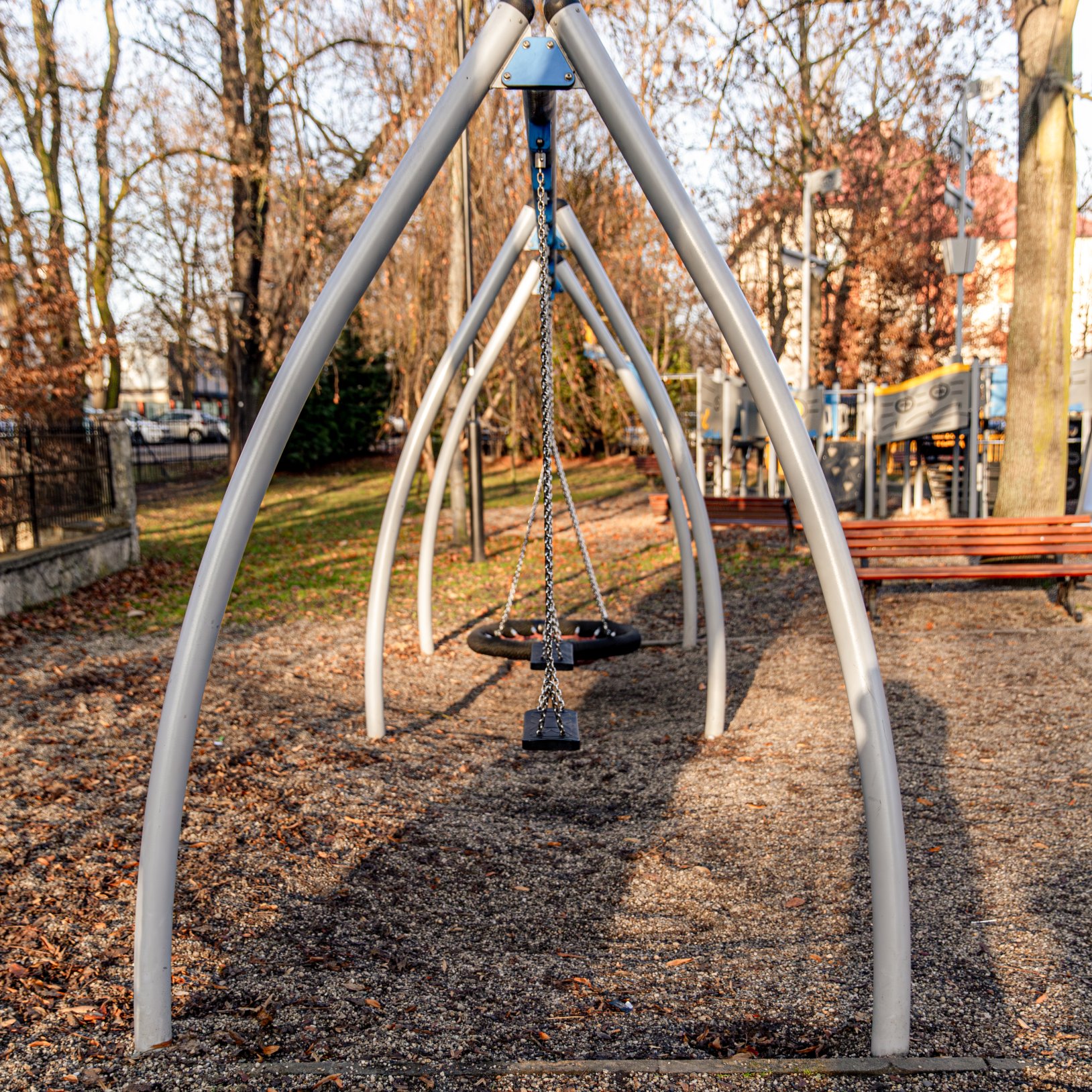 "Plac zabaw z nowoczesną huśtawką zawieszoną na metalowej konstrukcji w kształcie łuków. Huśtawka posiada dwa różne siedziska. Otoczenie stanowią drzewa bez liści, ławki oraz inna infrastruktura placu zabaw. Podłoże wysypane drobnym żwirem lub korą."