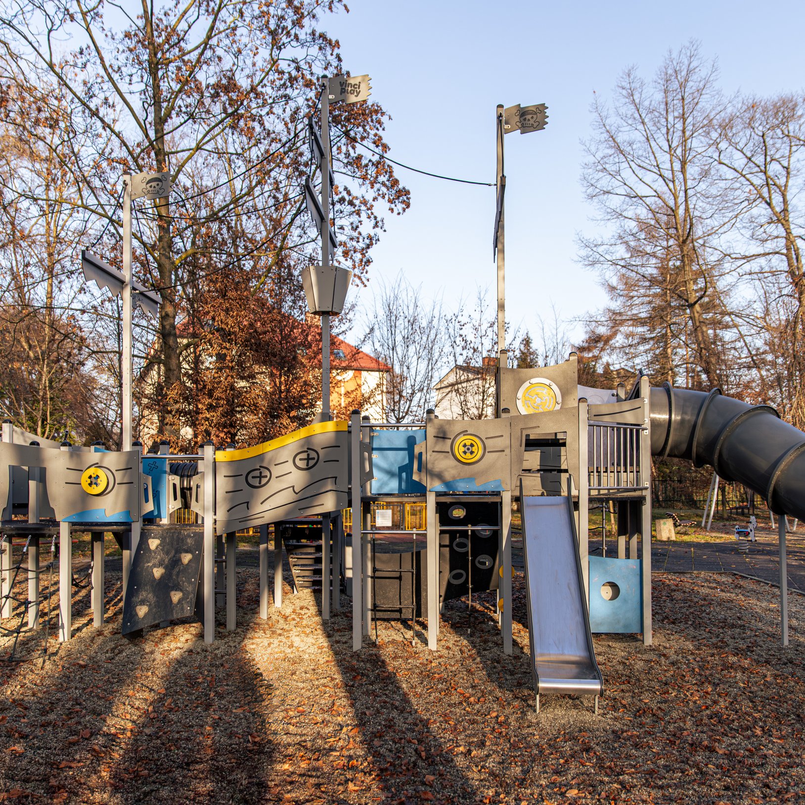 "Rozbudowana konstrukcja placu zabaw w formie statku lub pojazdu, z licznymi zjeżdżalniami, wspinaczkami i panelami edukacyjnymi. Dominują kolory szary, niebieski i żółty. W tle drzewa bez liści i zabudowania mieszkalne. Na ziemi widoczna warstwa kory drz