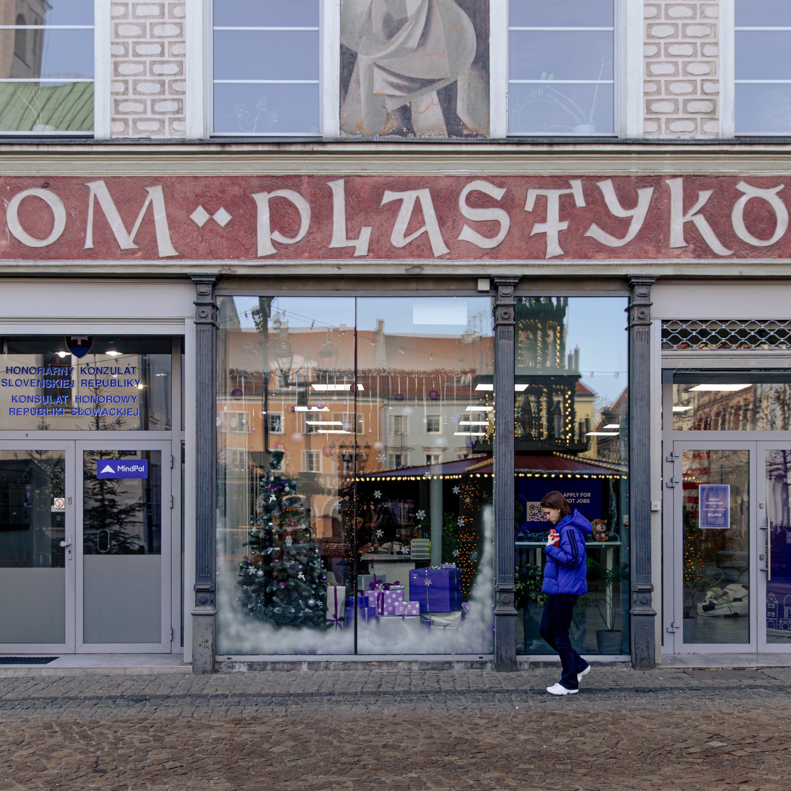 "Front budynku z czerwonym szyldem 'Dom Plastyków'. Na parterze znajdują się przeszklone witryny z dekoracjami świątecznymi oraz drzwi z napisem 'Konsulat Honorowy Republiki Słowackiej'. Przed budynkiem przechodzi osoba w niebieskiej kurtce."