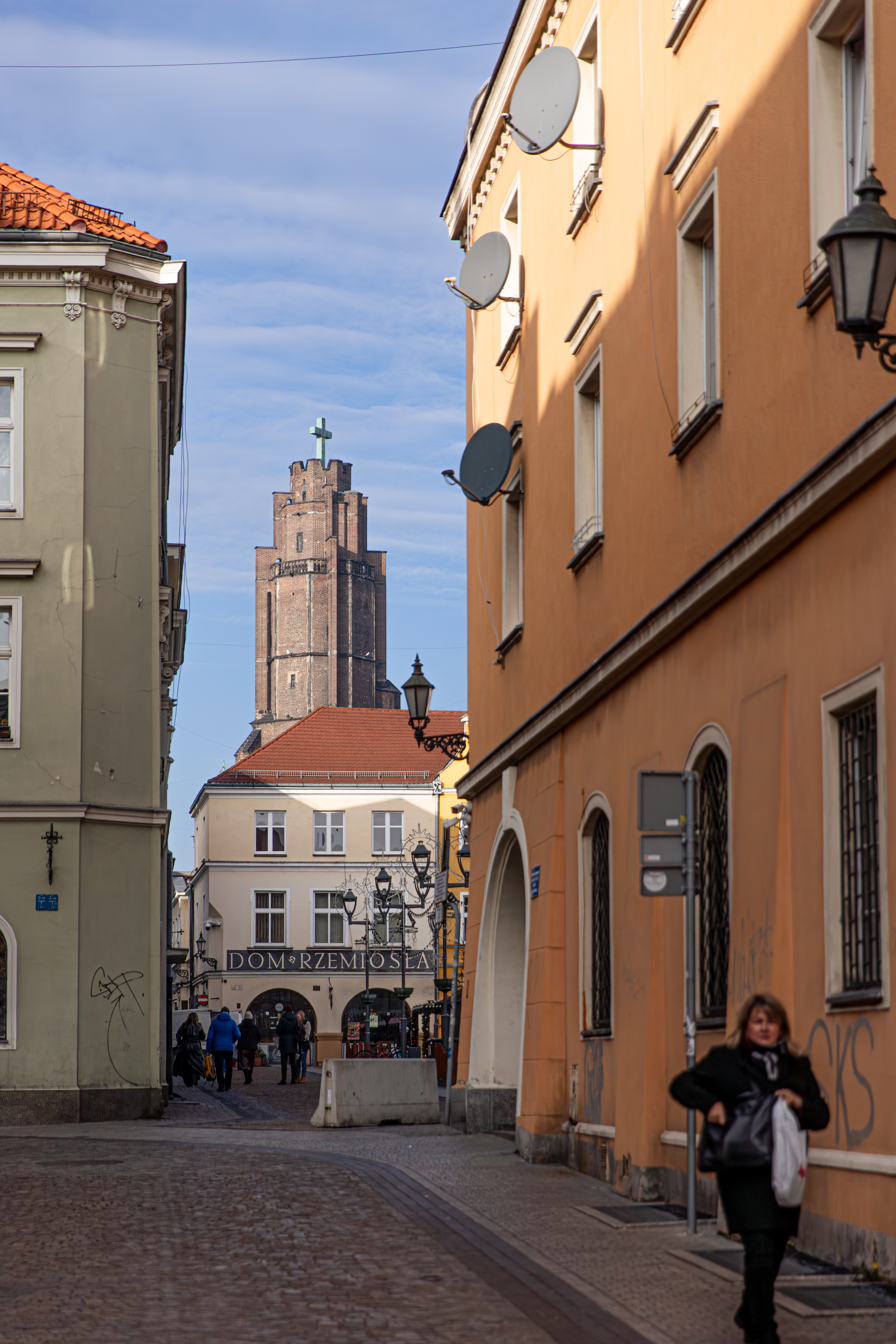 "Ulica prowadząca do budynku 'Dom Rzemiosła', widocznego w tle. Po bokach ulicy znajdują się kamienice, jedna w kolorze oliwkowym, druga pomarańczowa z licznymi antenami satelitarnymi. W tle widać wieżę kościoła z krzyżem. Na ulicy znajdują się piesi."
