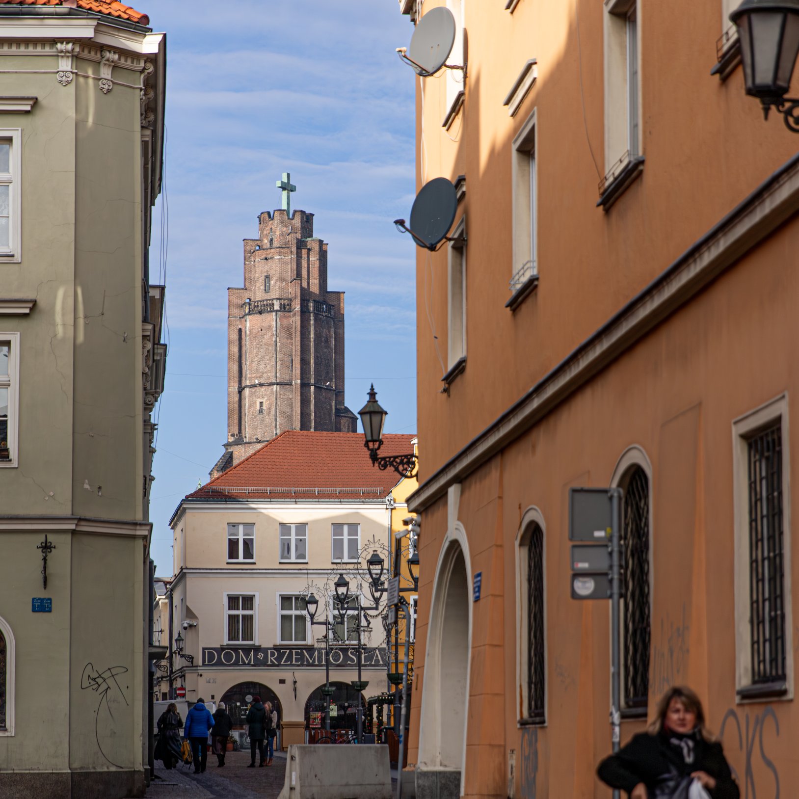 "Ulica prowadząca do budynku 'Dom Rzemiosła', widocznego w tle. Po bokach ulicy znajdują się kamienice, jedna w kolorze oliwkowym, druga pomarańczowa z licznymi antenami satelitarnymi. W tle widać wieżę kościoła z krzyżem. Na ulicy znajdują się piesi."