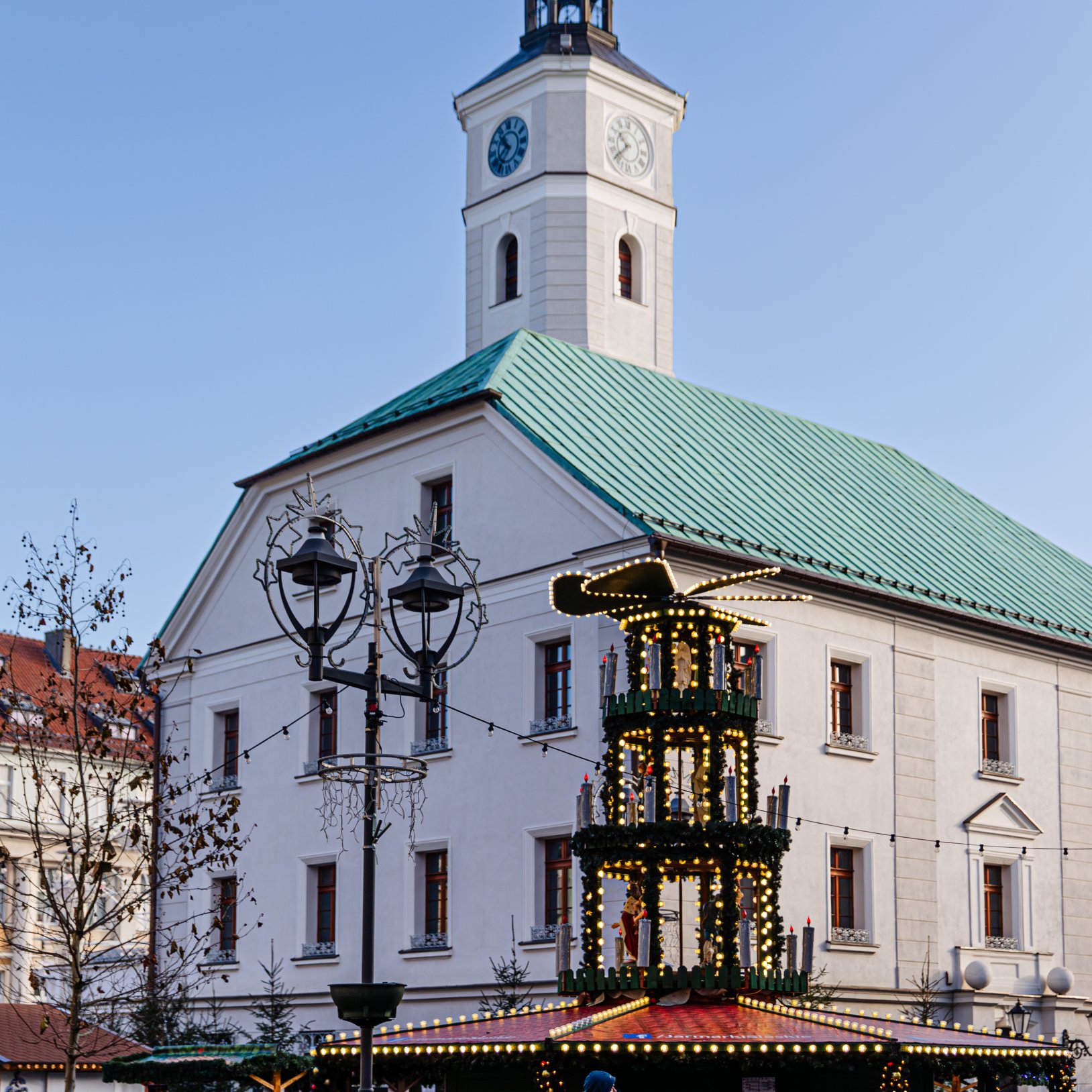 "Widok rynku z ozdobnym drewnianym kioskiem świątecznym udekorowanym girlandami i światełkami. W tle znajduje się biały budynek z zielonym dachem i wieżą zegarową. Na pierwszym planie widać przechodnia i latarnie miejskie."