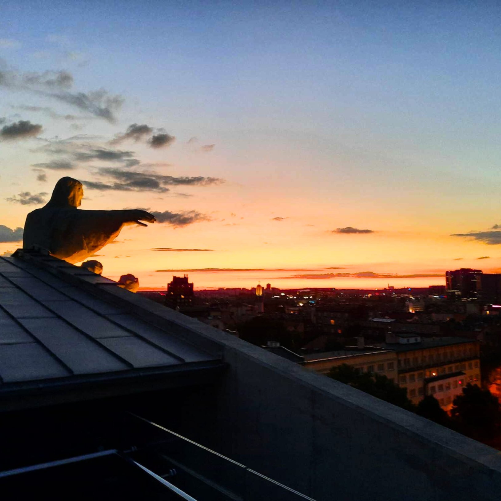 "Widok z wysokości na panoramę miasta o zmierzchu. Na pierwszym planie widać rzeźbę postaci z rozpostartym ramieniem na dachu budynku. W tle rozciąga się miasto z oświetlonymi budynkami i wieżowcami, a niebo mieni się barwami zachodu słońca – od pomarańcz